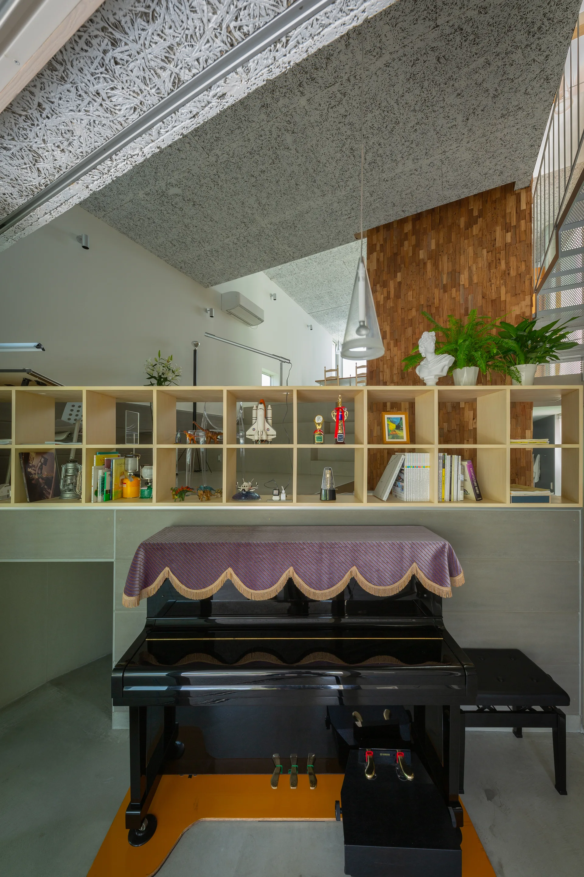 A black upright piano sits below a light wood cube bookshelf against a concrete wall and wood slat paneling in a contemporary space.