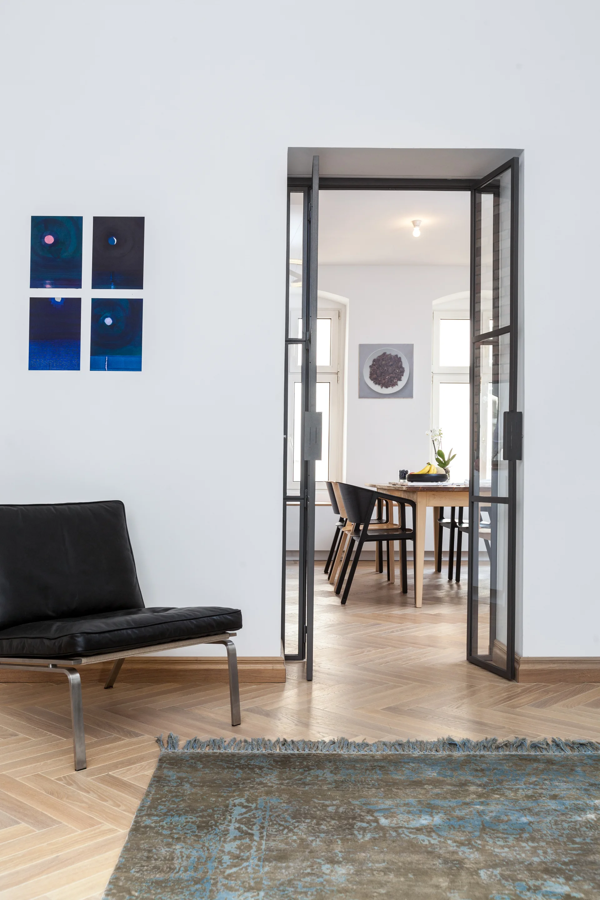 A black metal-framed glass doorway opens from a living room with a leather chair onto a dining room with a wooden table and herringbone floors.
