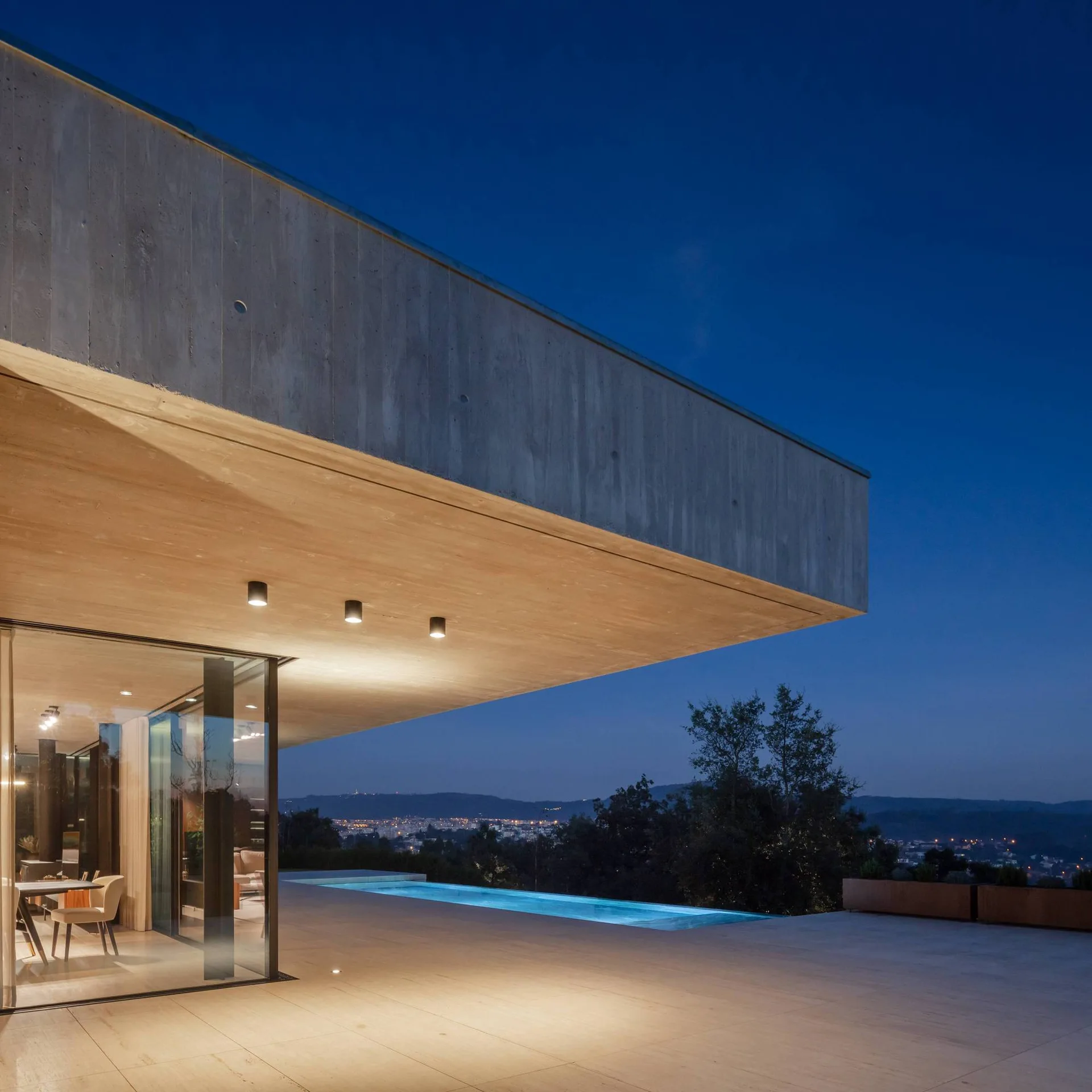 A modern patio with a concrete cantilevered overhang, recessed spotlights, a large tiled terrace, and an outdoor swimming pool at twilight.