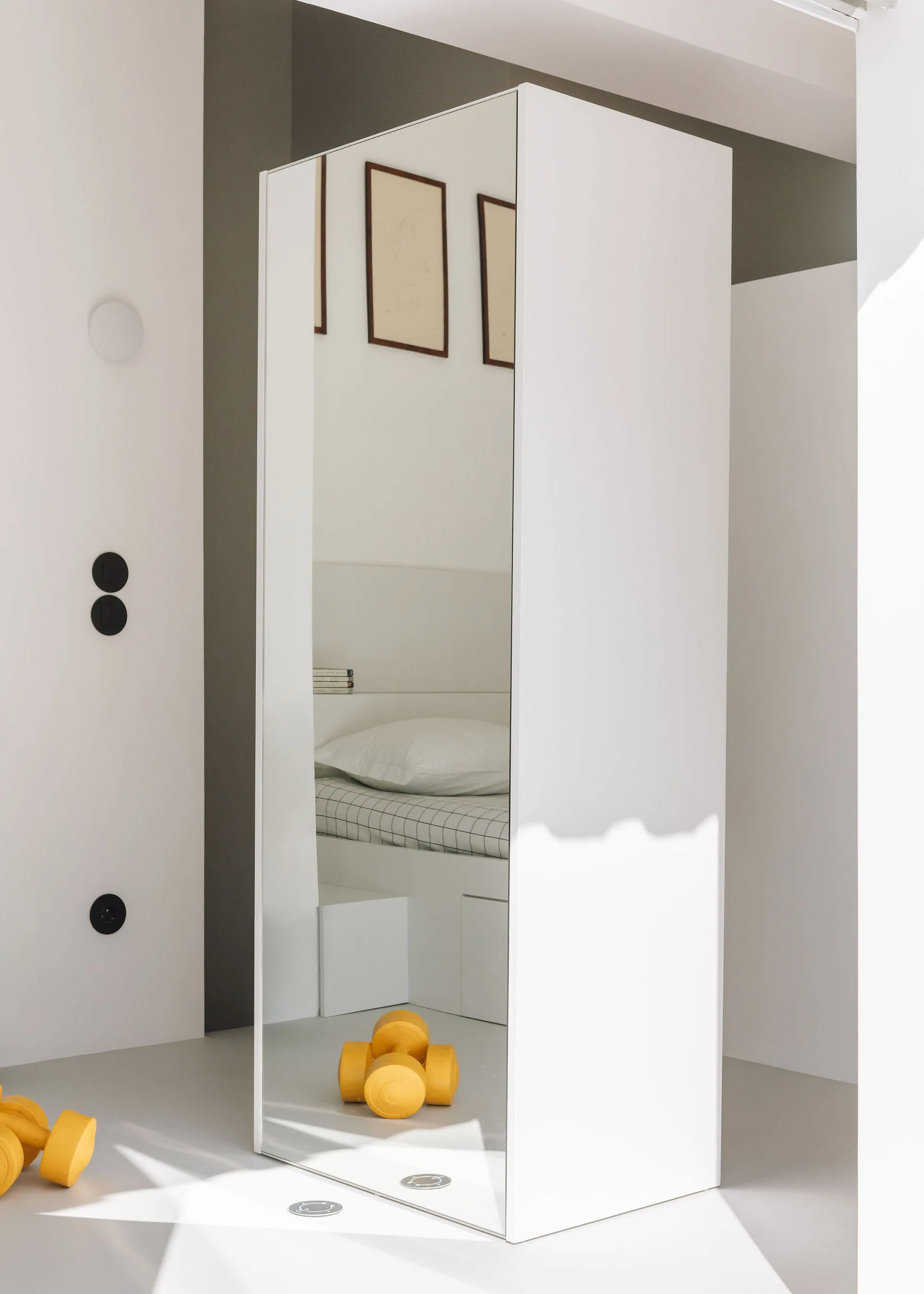 A minimalist white bedroom with a full-length mirrored wardrobe reflecting a bed with grid-patterned bedding and yellow dumbbells on the grey floor.