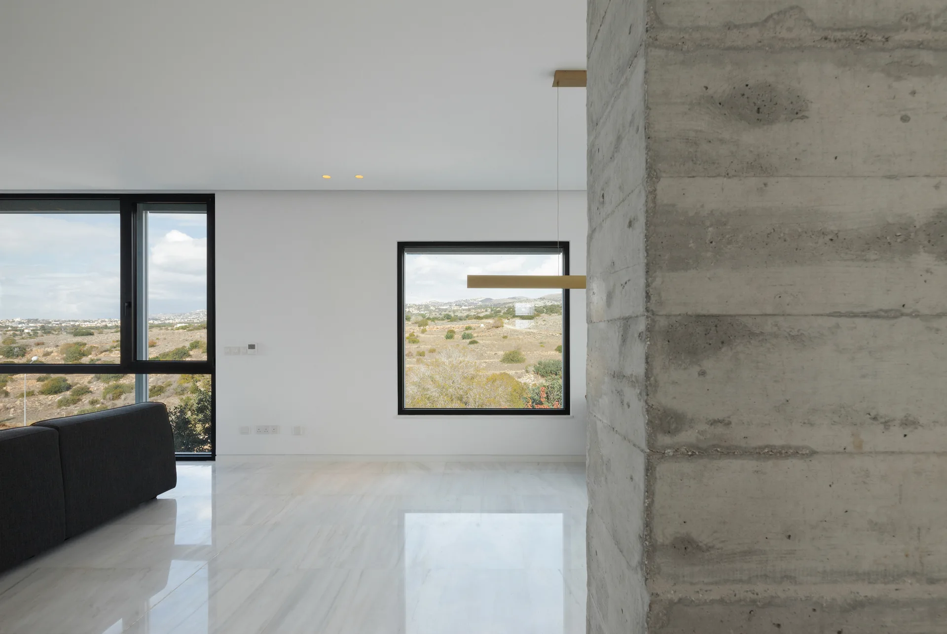 A minimalist living room features a textured concrete pillar, polished stone floors, and large black-framed windows overlooking a landscape.