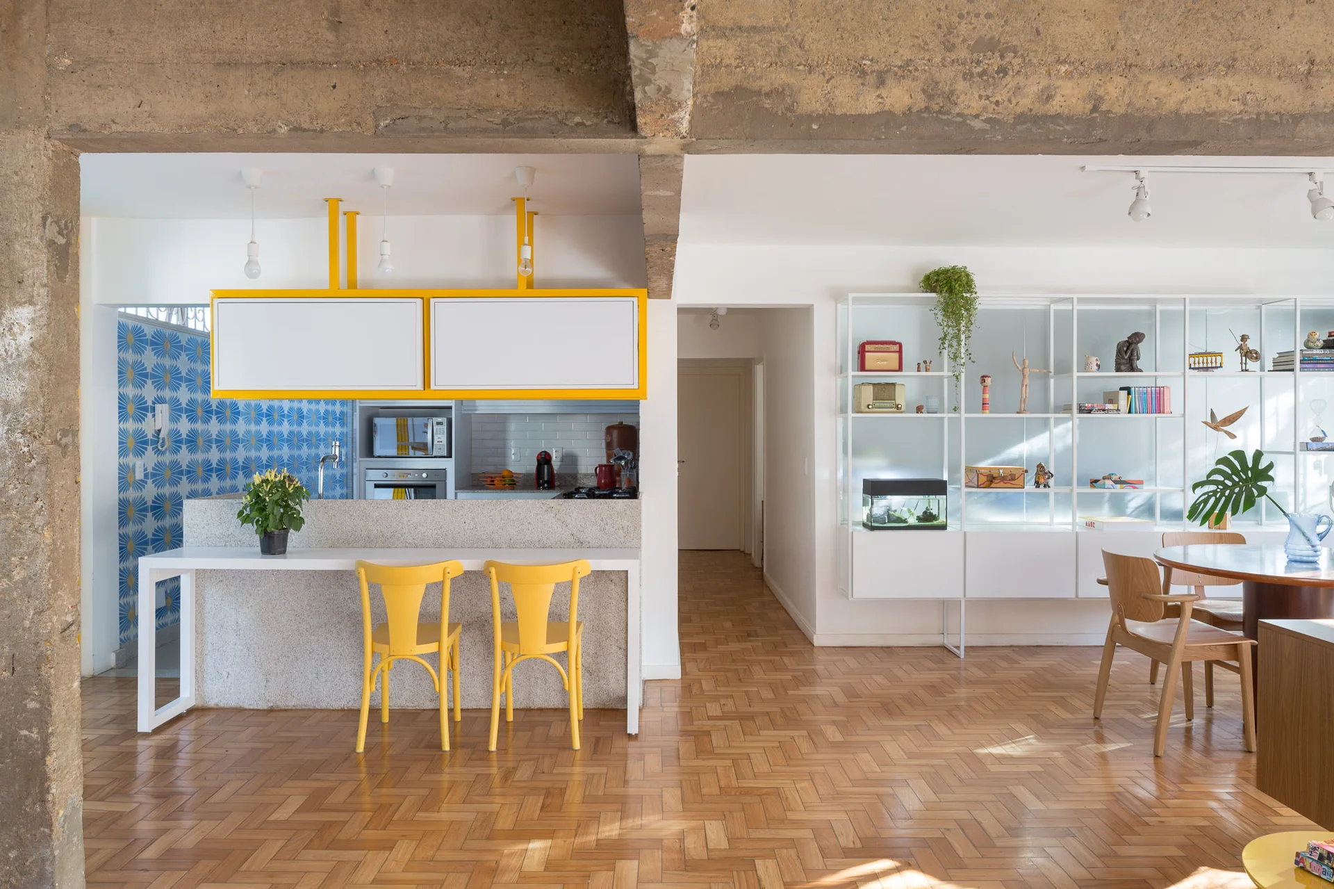 An open kitchen features yellow bar stools, suspended cabinets with yellow trim, a stone island, and exposed concrete ceiling beams.