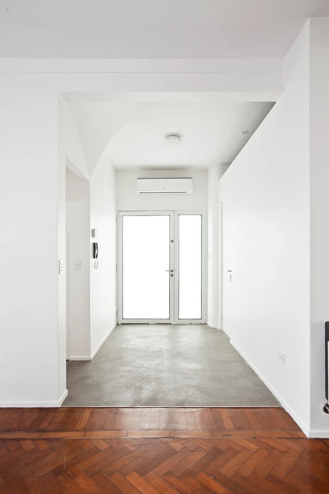 A minimalist white entryway featuring a transition from rich wood herringbone parquet flooring to a smooth grey concrete floor leading to a glass front door.