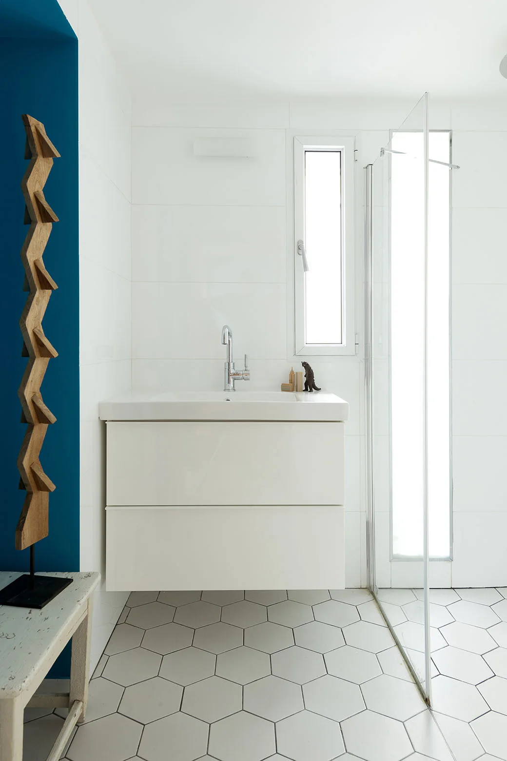 A modern bathroom features a white wall-mounted vanity, a glass shower screen, white hexagonal floor tiles, and a bold blue accent wall on the left.