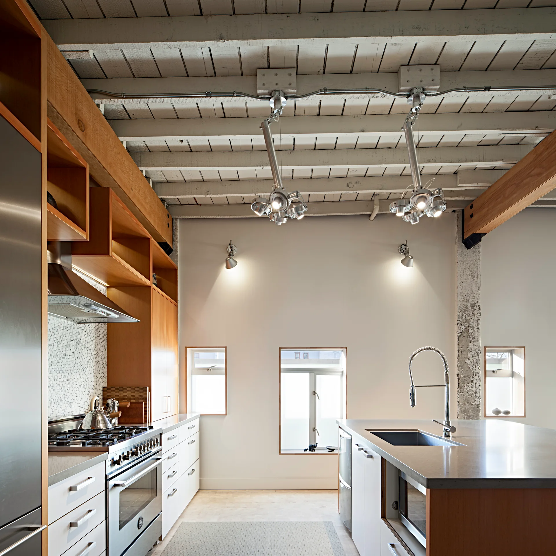 An industrial loft kitchen features a stainless steel range, white base cabinets, a central island, and exposed white-painted wood ceiling beams.