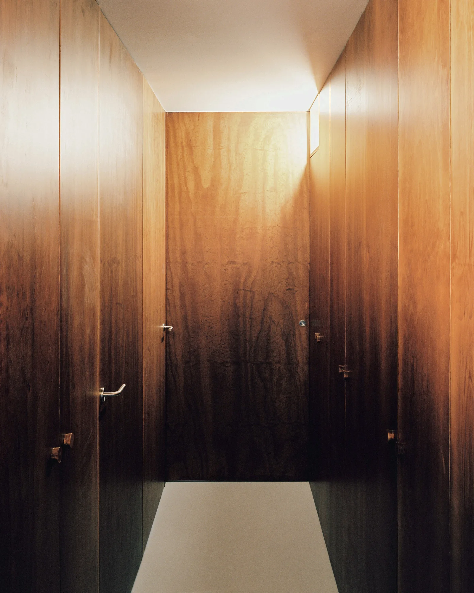 A narrow minimalist hallway features floor-to-ceiling warm wood paneling, simple metal door handles, a white floor, and a white ceiling.