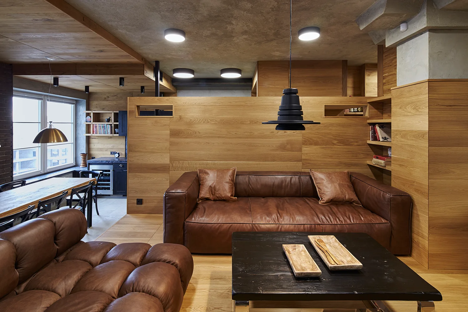 A modern living room features brown leather sofas, a black coffee table, wood-paneled walls, and a textured concrete ceiling with recessed lights.