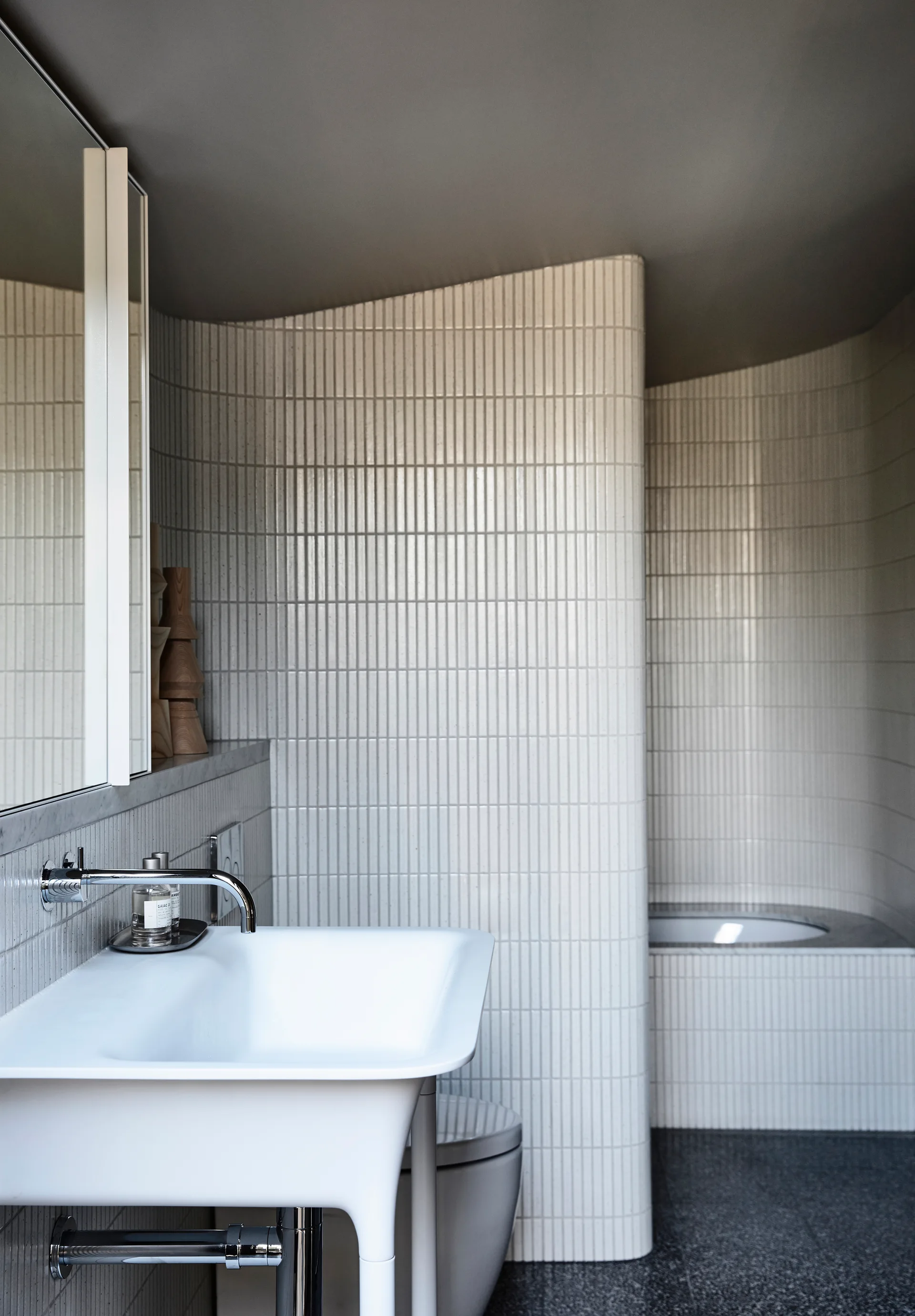 A modern bathroom with a white wall-mounted basin, a chrome faucet, and a curved wall covered in slender vertical white tiles.