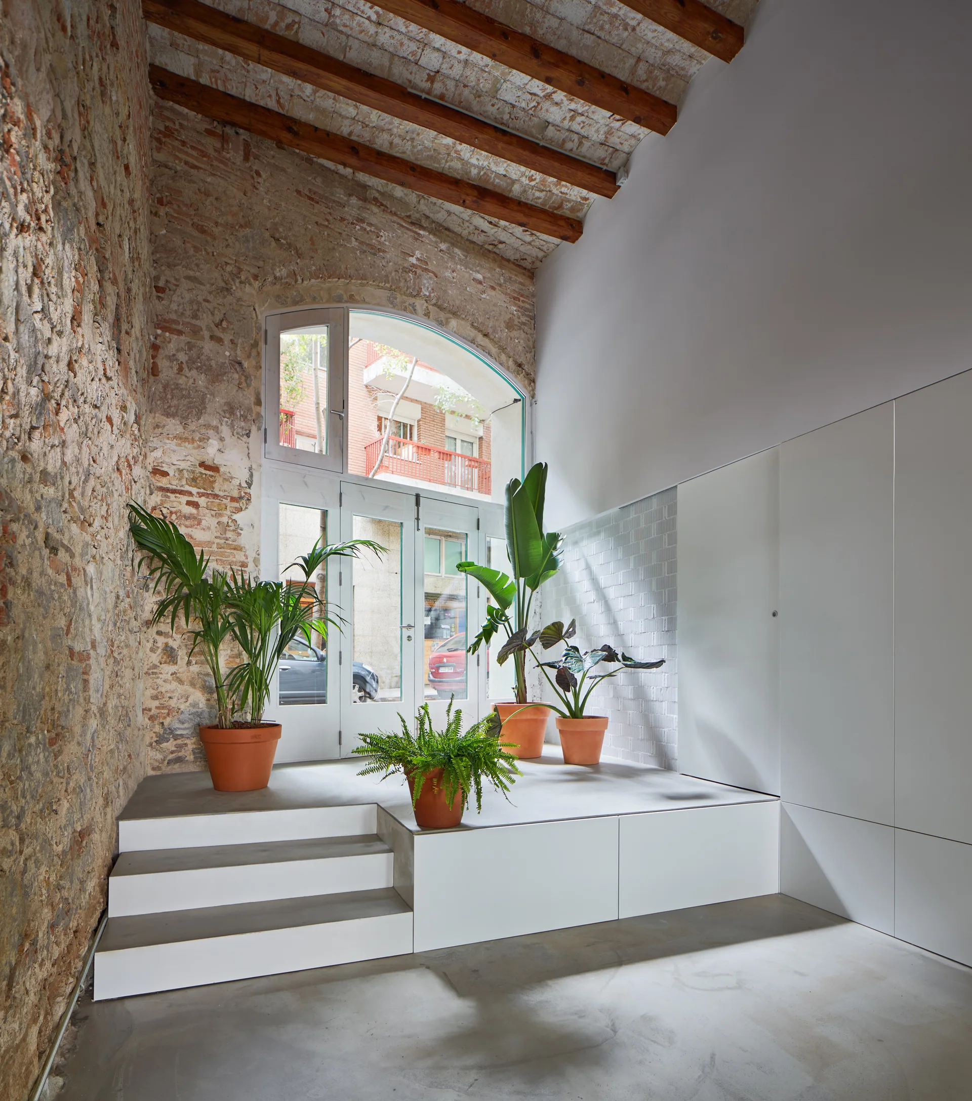 A bright entryway features a white stepped platform with potted plants next to an exposed brick wall and a large arched glass door.