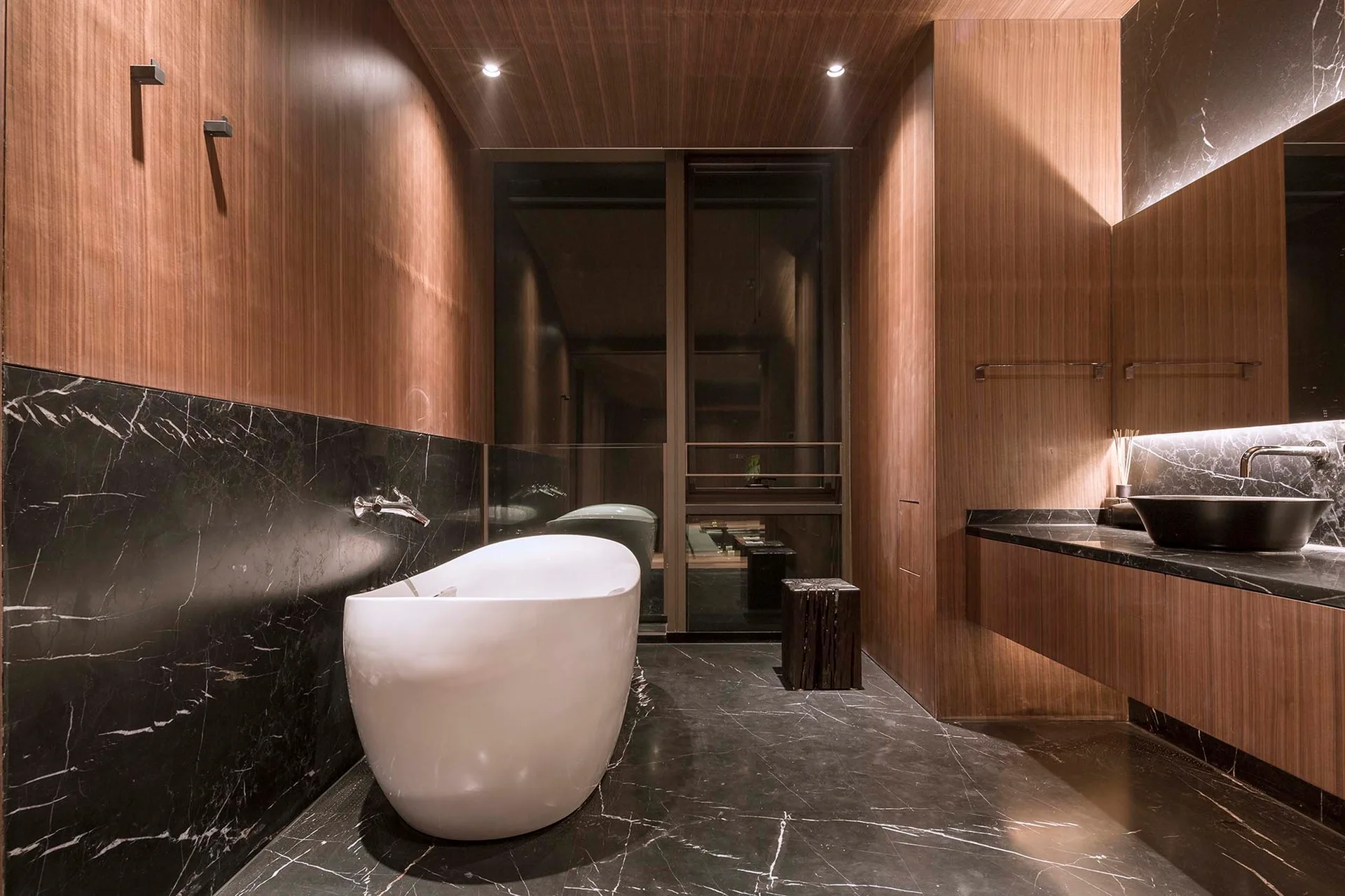 A modern bathroom featuring a white freestanding bathtub, black veined marble floors, and vertical wood paneling on the walls and ceiling.