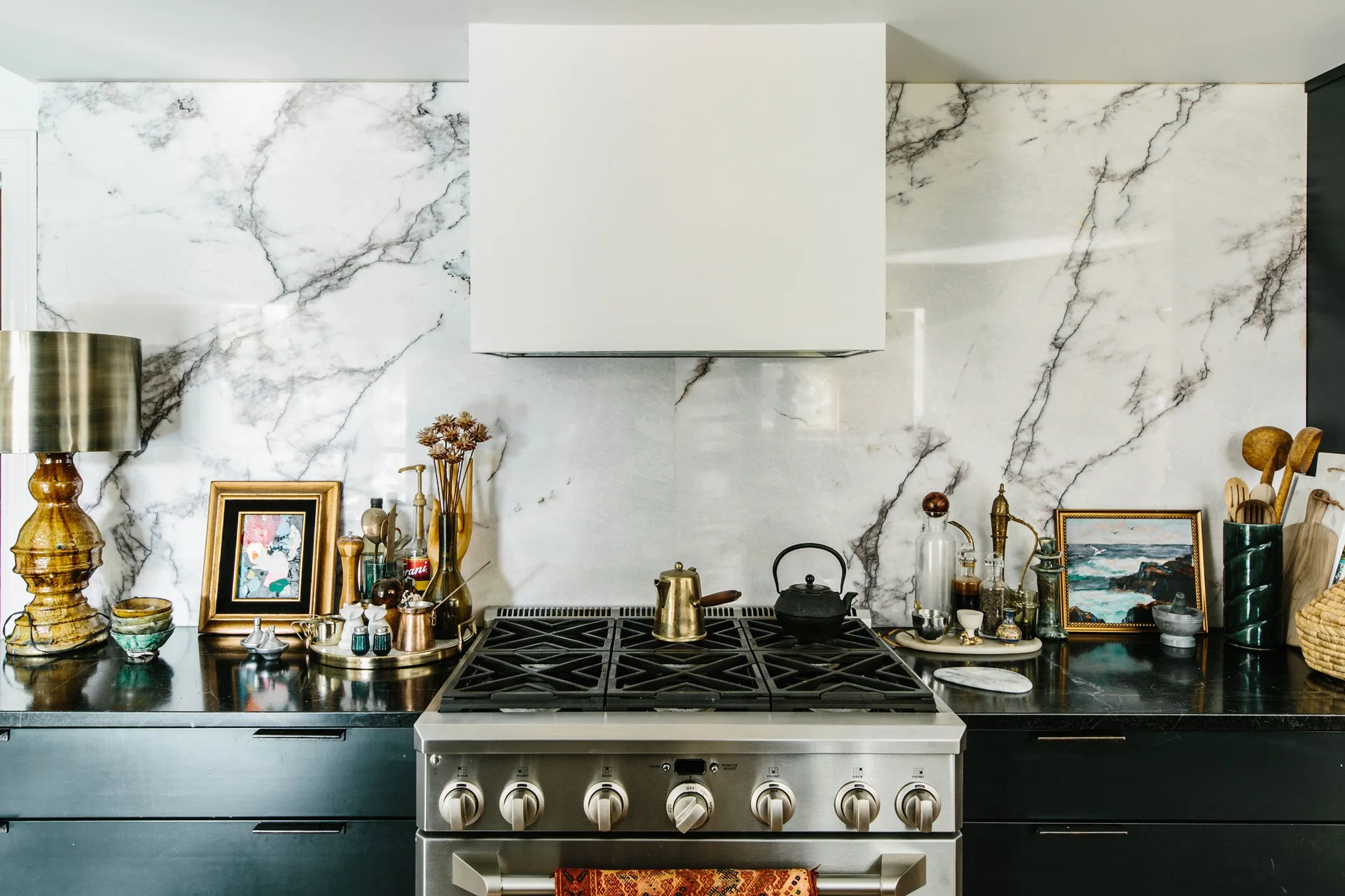 A stainless steel kitchen range with a white hood and a veined marble backsplash, flanked by black countertops styled with framed art and small decor.