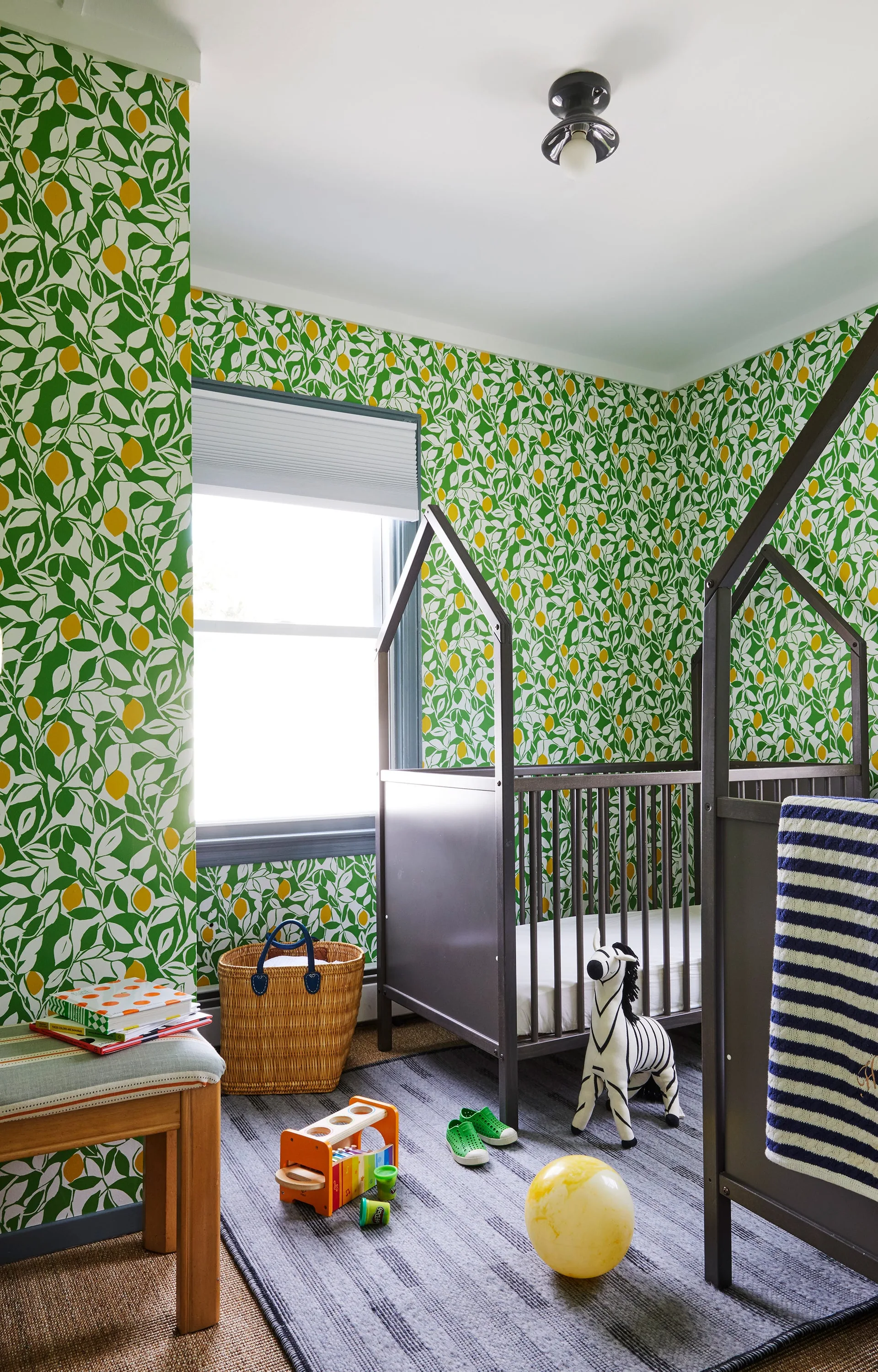 A nursery featuring green and yellow lemon-patterned wallpaper, a dark grey house-frame crib, a striped rug, and a small woven basket.
