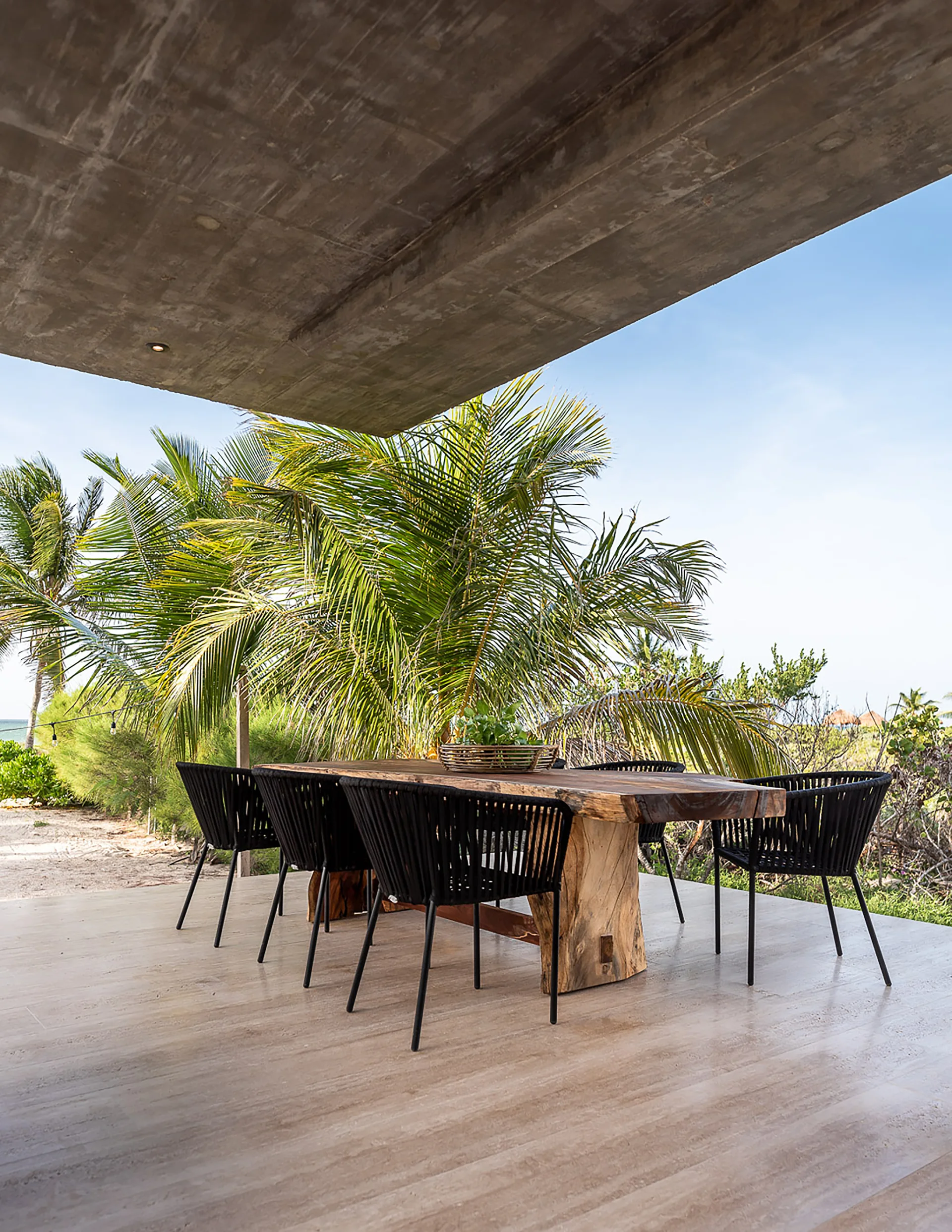 An outdoor patio dining area features a large live-edge wood table and black woven chairs under a massive concrete ceiling overlooking palm trees.