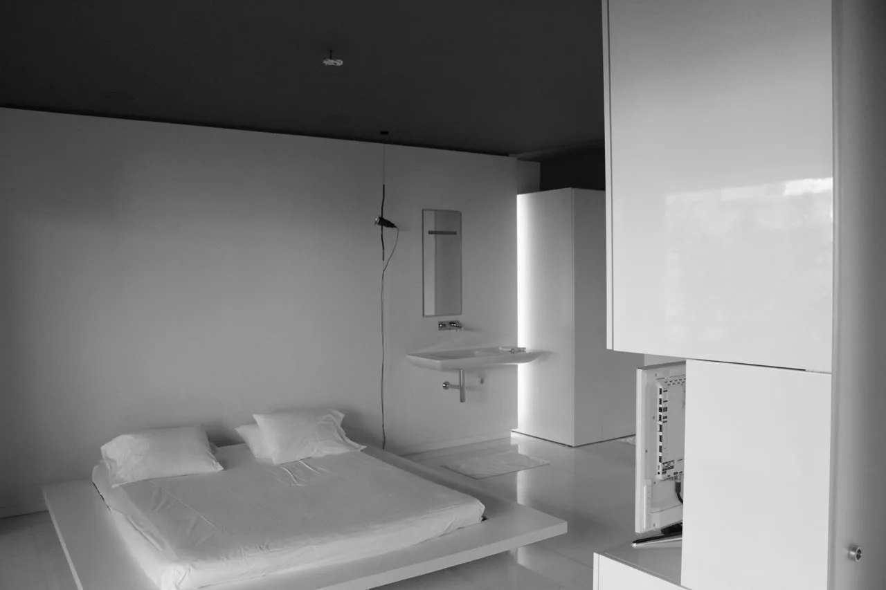 A minimalist bedroom features a low white platform bed, glossy floors, a dark ceiling, and a wall-mounted sink with a slim mirror.