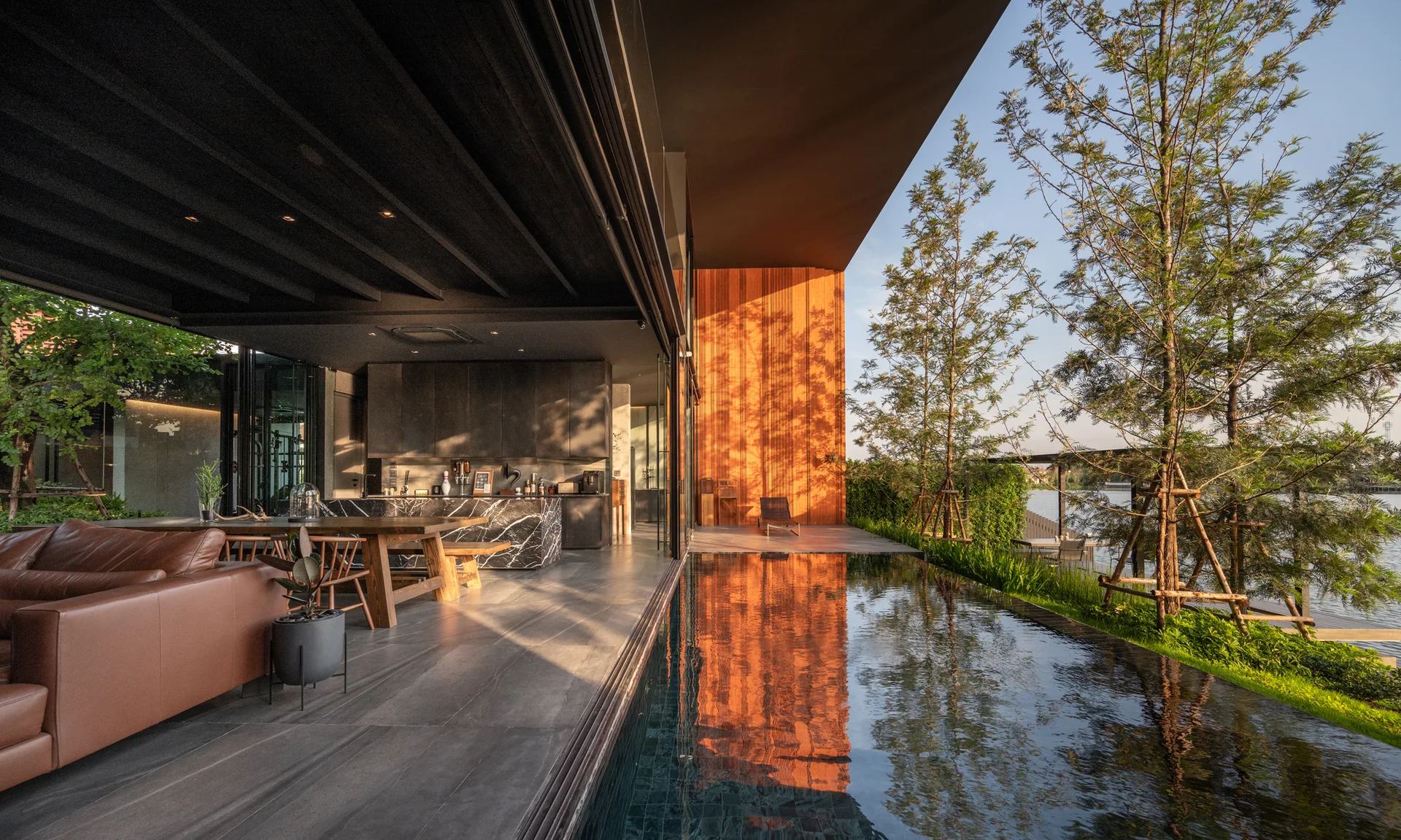 A modern indoor-outdoor living room and kitchen featuring a tan leather sofa, black marble island, and a swimming pool reflecting a warm wood wall.