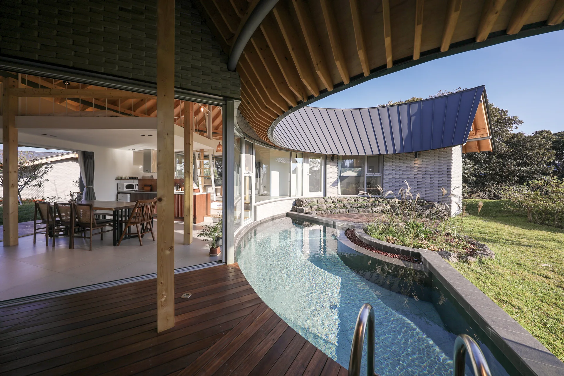 A modern curved house exterior features a integrated swimming pool, wooden decking, and a dining area visible through open sliding glass doors.