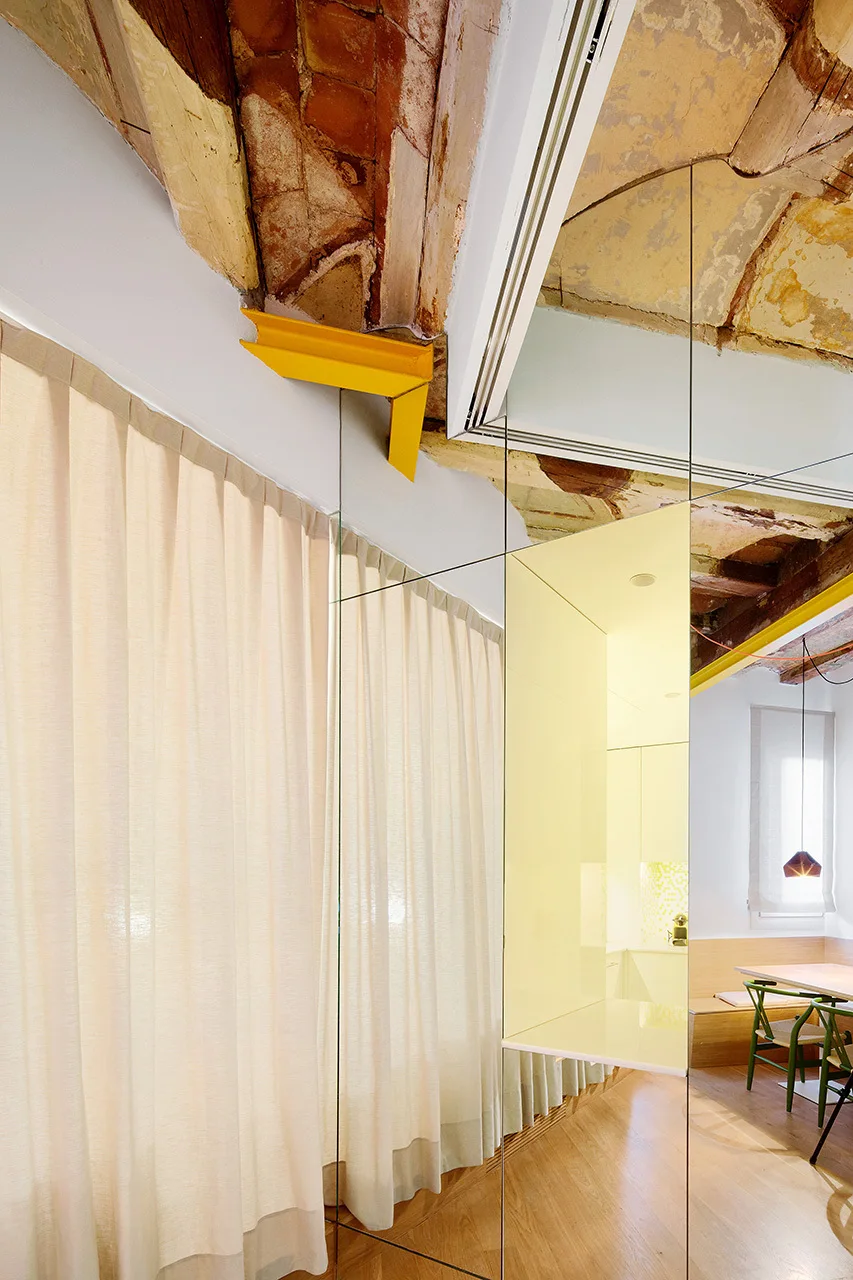 A dining space features an exposed brick vaulted ceiling, a large mirrored wall reflecting a dining set, and a bright yellow structural beam.
