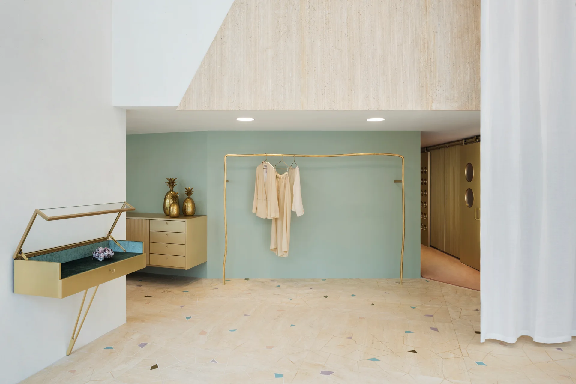 A dressing room featuring a wavy gold clothing rack against a sage green wall, a wall-mounted dresser, and light terrazzo flooring.