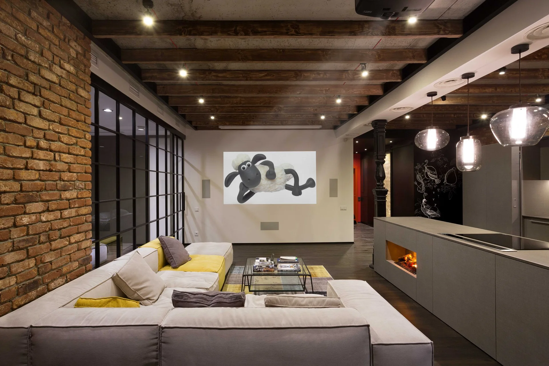 An industrial loft living space featuring a modular sofa, a brick accent wall, wooden ceiling beams, and a kitchen island with an integrated fireplace.
