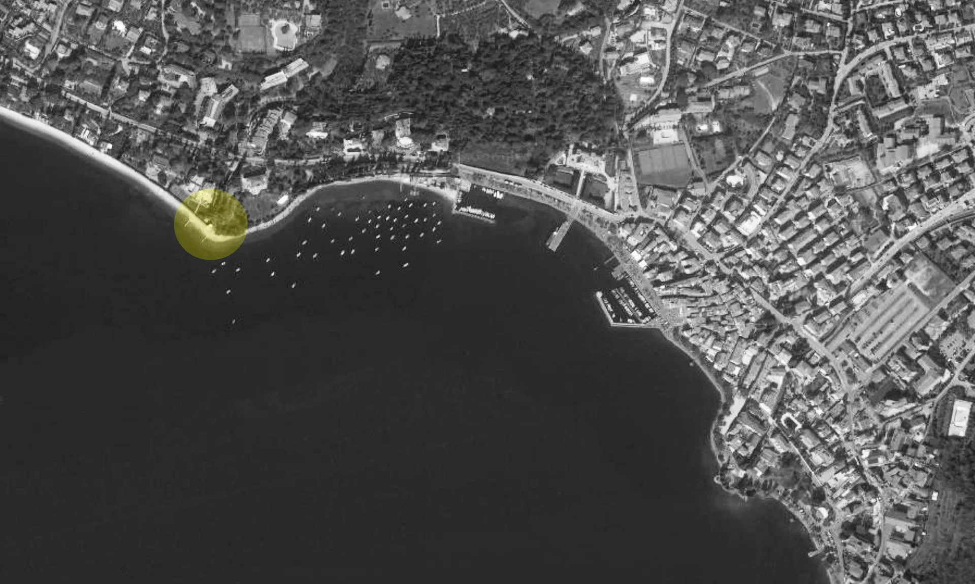 A black and white aerial satellite map of a coastal town and marina, with a translucent yellow circle highlighting a specific plot along the shoreline.