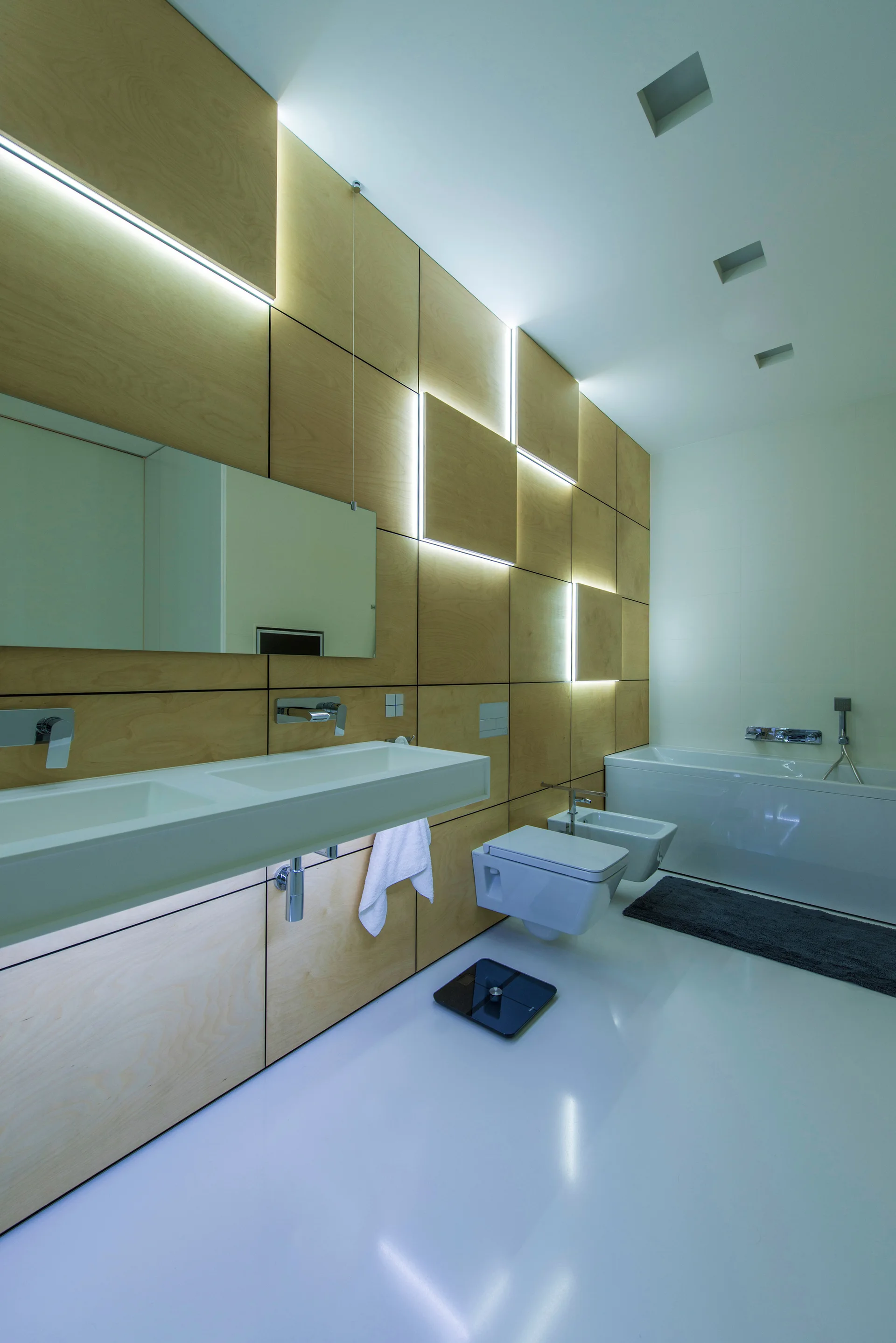 A contemporary bathroom features a light wood paneled wall with integrated backlighting, a white double vanity, and wall-hung toilet fixtures.