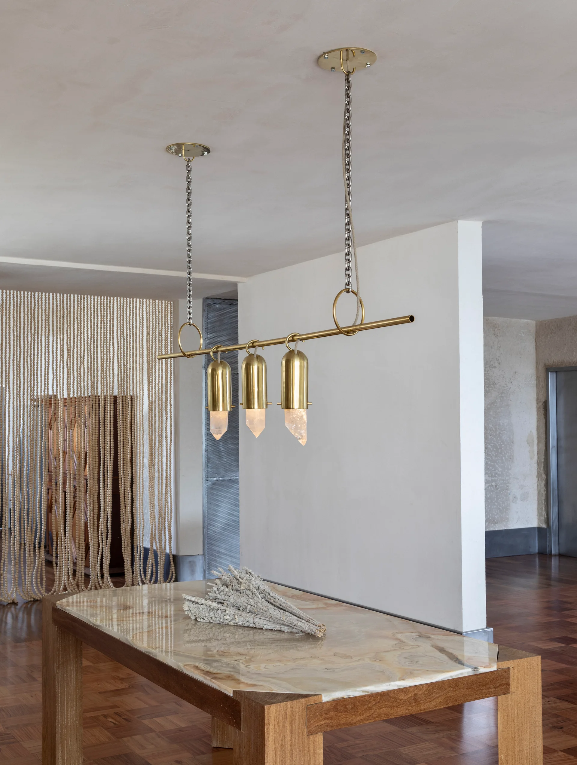 A brass pendant light with quartz crystals hangs above a stone-topped wooden table in a room with a beaded curtain in the background.
