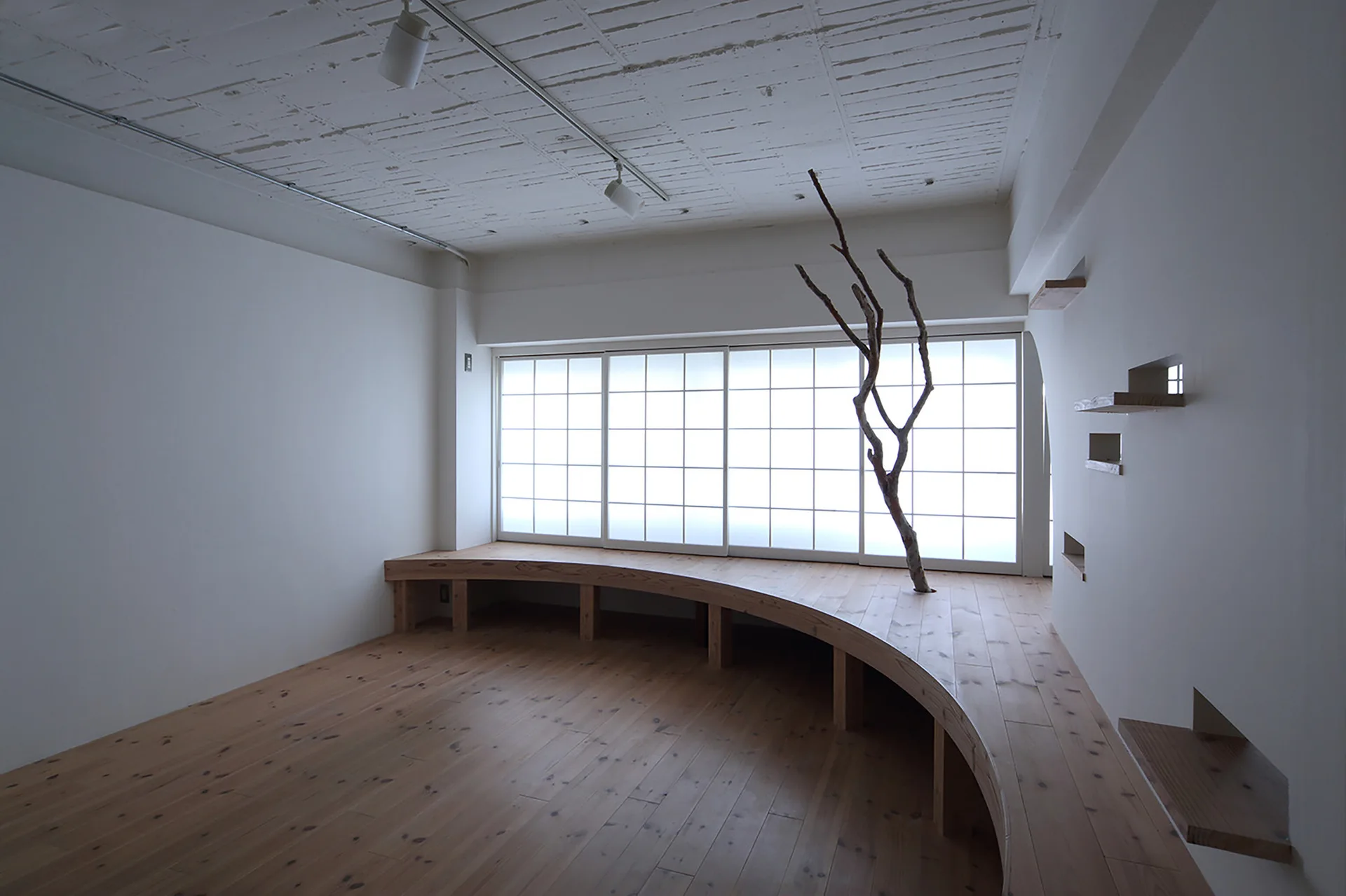 A minimalist interior features a curved wooden platform bench beneath a large gridded window with a single decorative dry branch.