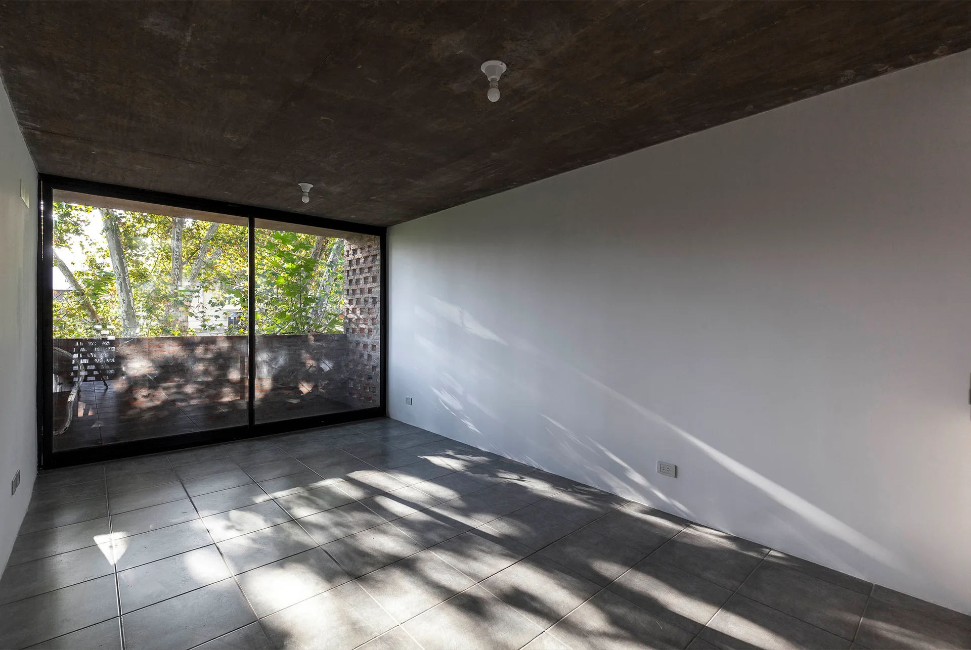 An empty minimalist room featuring an exposed concrete ceiling, plain white walls, grey tiled flooring, and a large sliding glass door opening to a balcony.