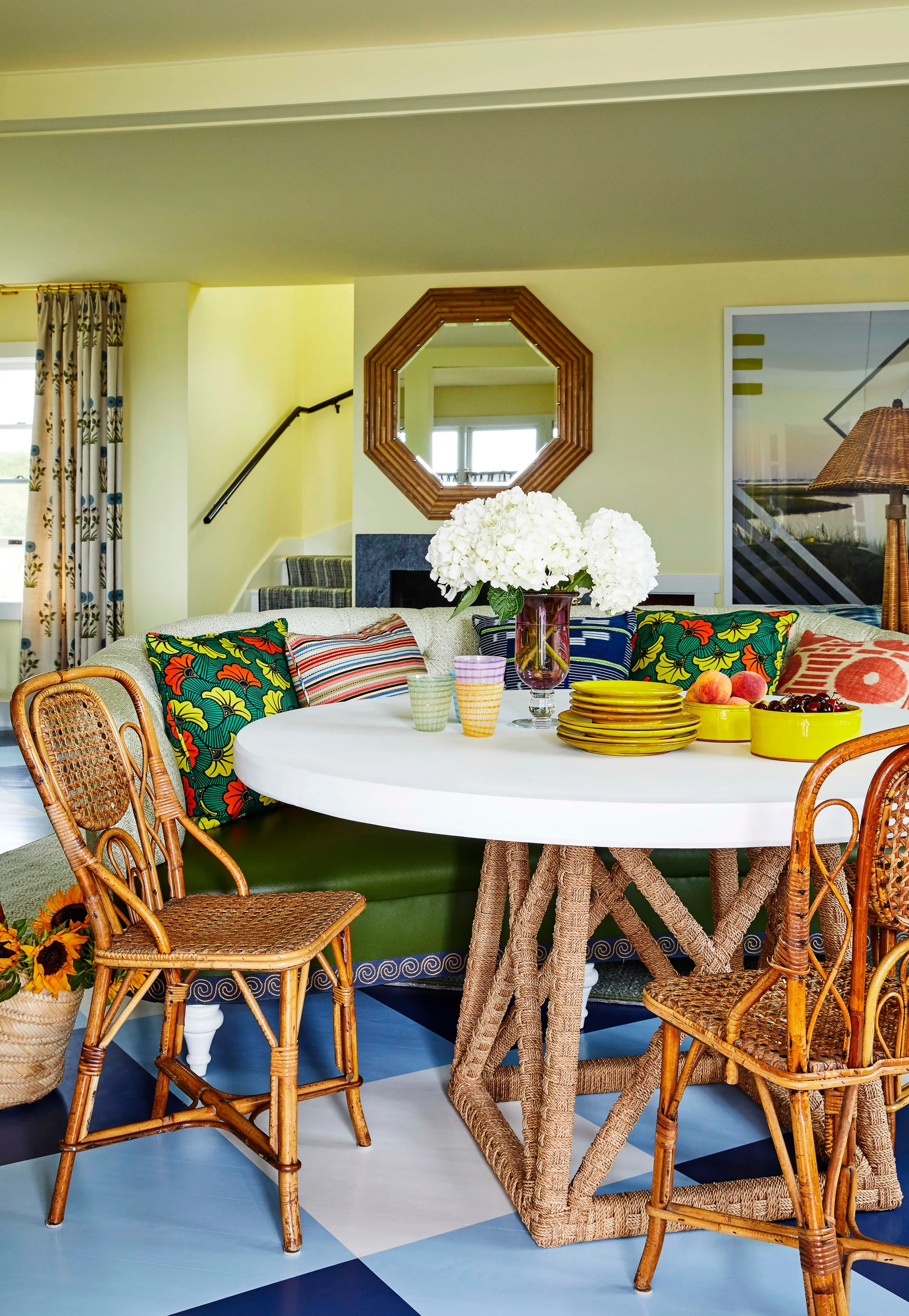A colorful breakfast nook features a round white table with a rope base, rattan chairs, and a curved banquette against pale yellow walls.