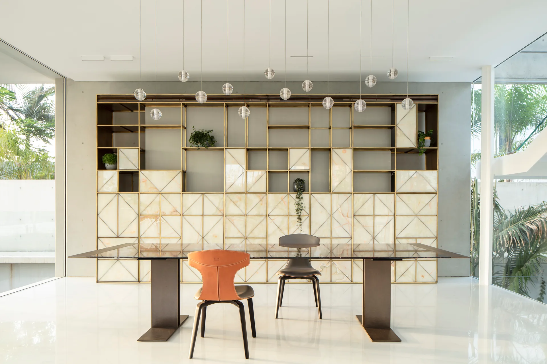 A modern dining room features a long glass table with chairs beneath a row of glass globe pendant lights and a large brass geometric shelving unit.