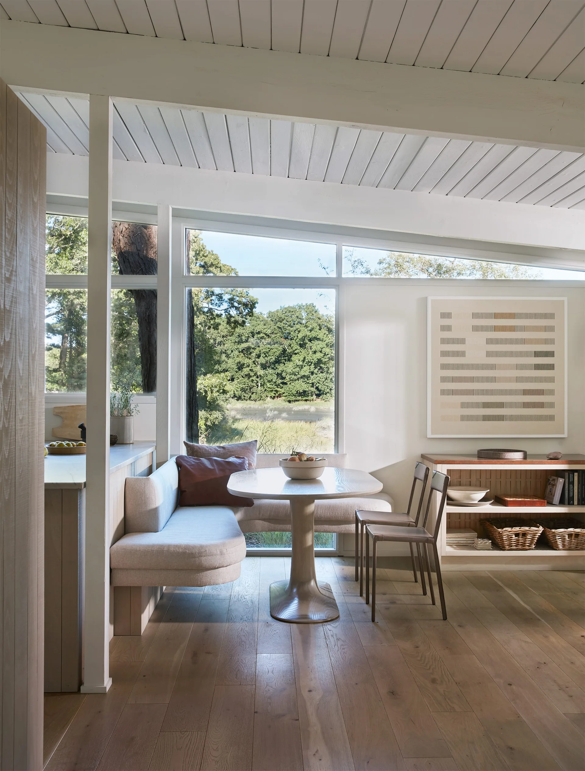A modern dining nook features a built-in beige banquette, a light wood pedestal table, and two chairs in front of a large picture window.