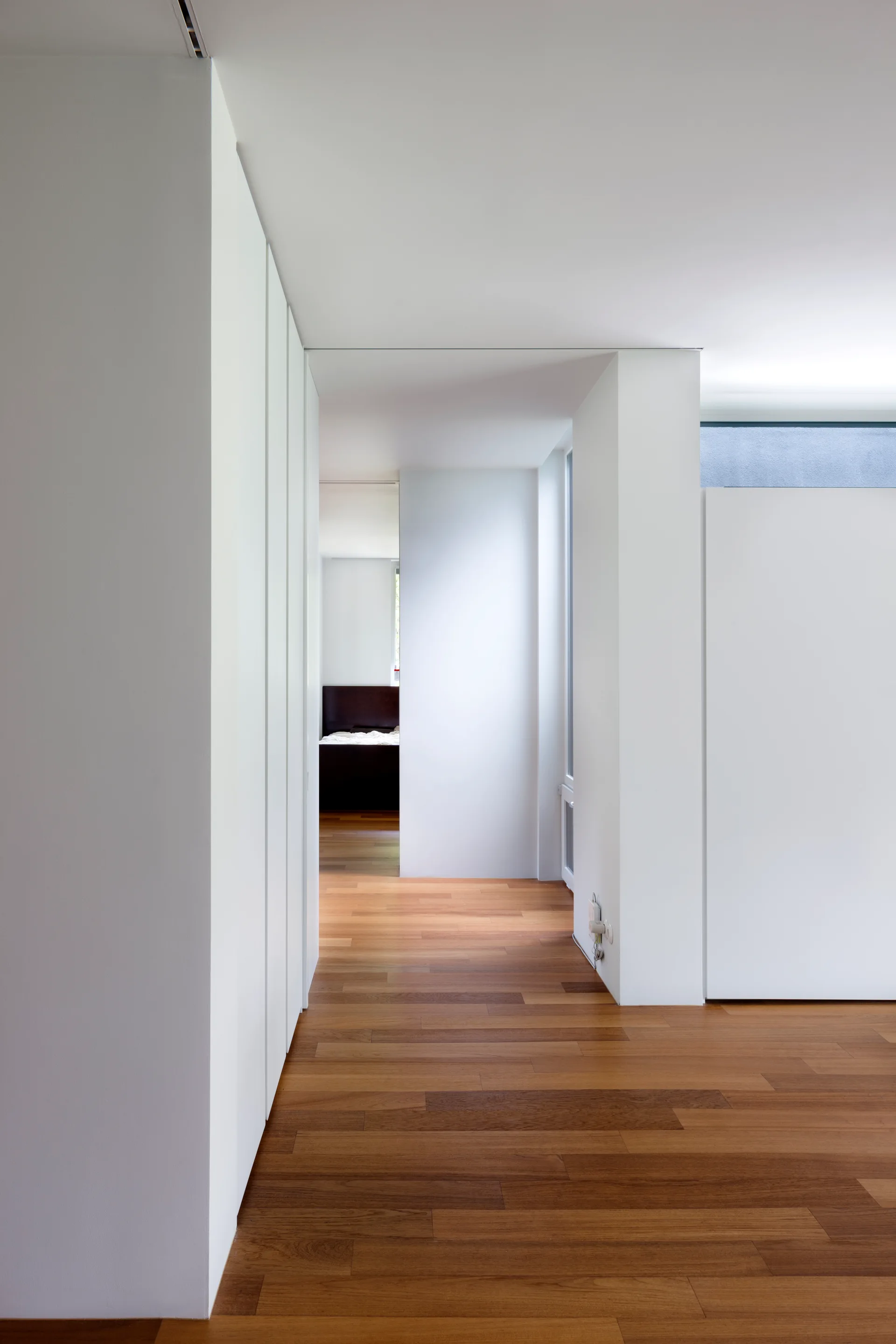 A clean white minimalist hallway features flush built-in closet doors on one side and warm hardwood plank flooring leading to a distant bedroom.