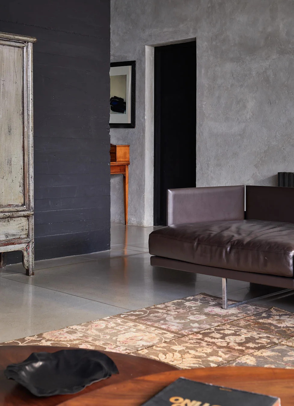 A brown leather sofa rests on polished concrete floors next to a textured grey plaster wall and a tall distressed wooden cabinet.