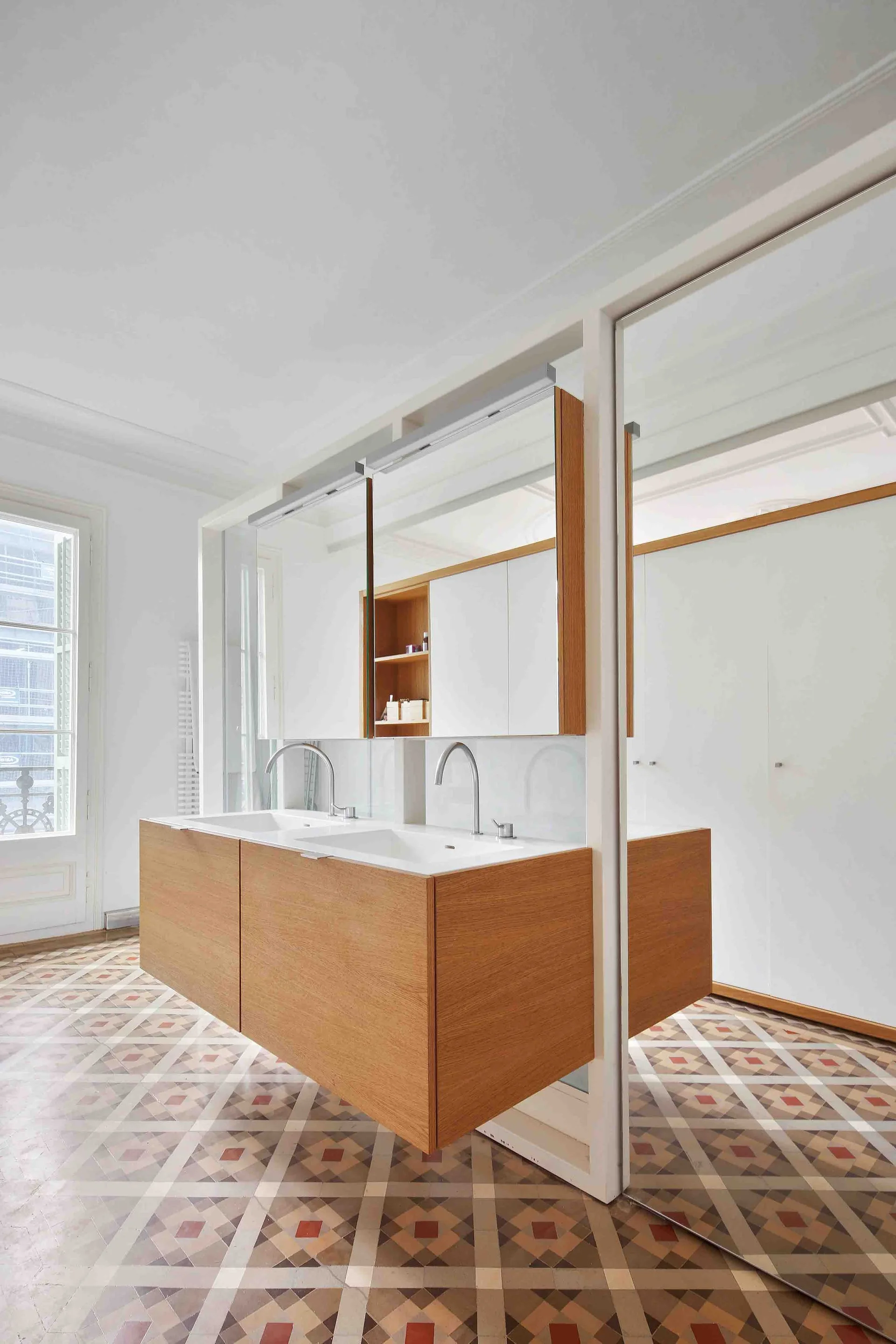 A floating double sink bathroom vanity with wood cabinetry sits against a partition wall on a geometric patterned tile floor.