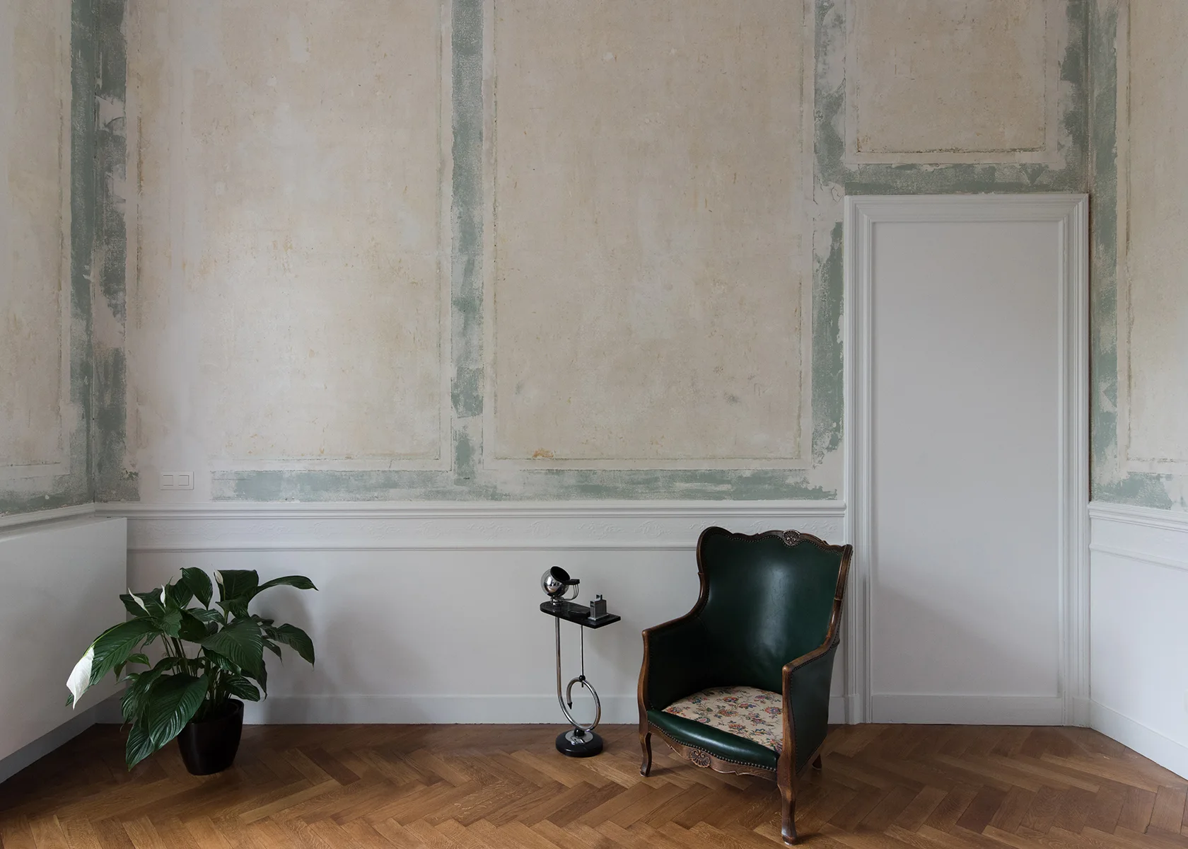A vintage green leather armchair and small side table sit on a parquet floor in a room with distressed plaster walls and white wainscoting.