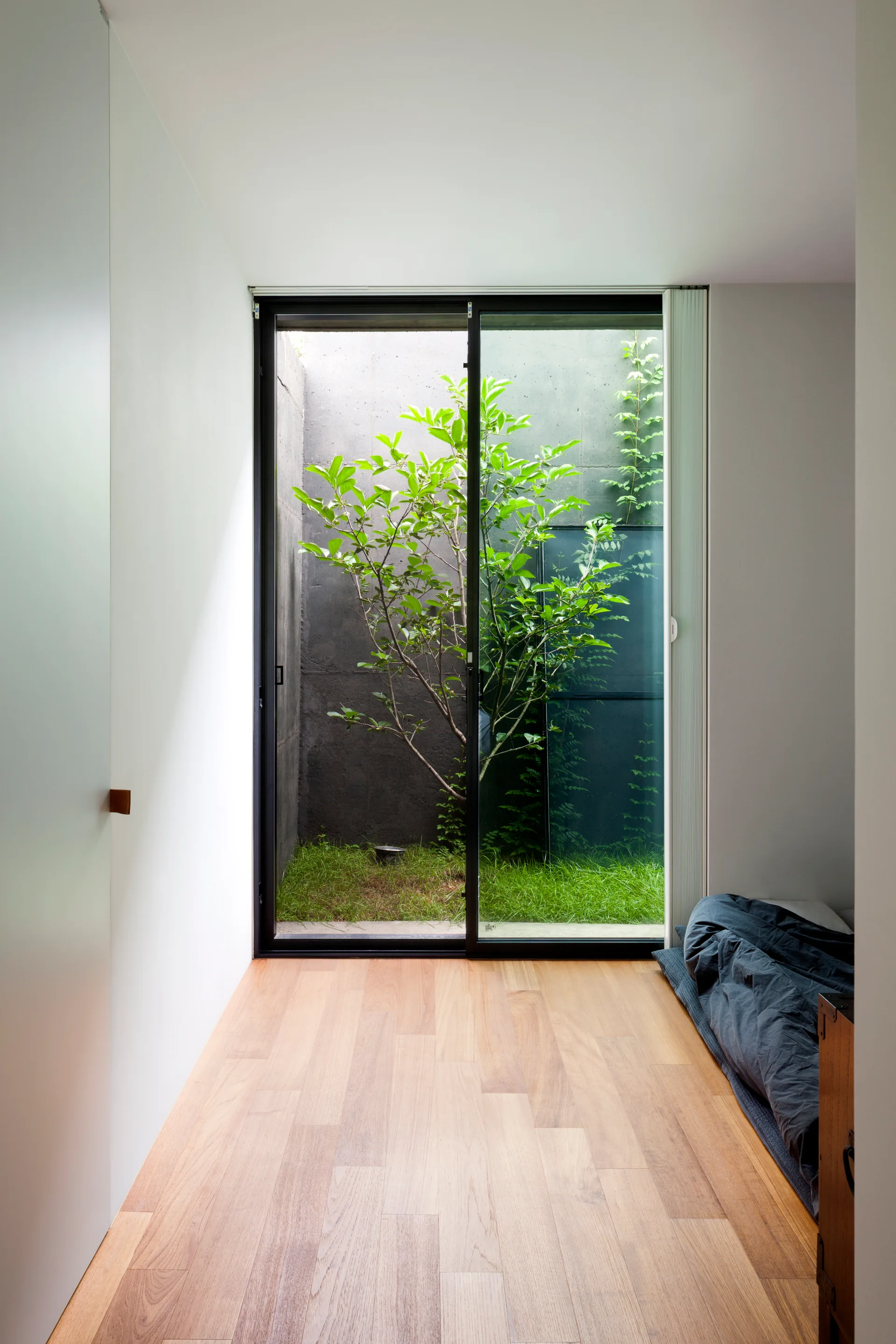 A minimalist bedroom featuring light wood floors and a low floor bed next to a large sliding glass door opening to a small garden courtyard.