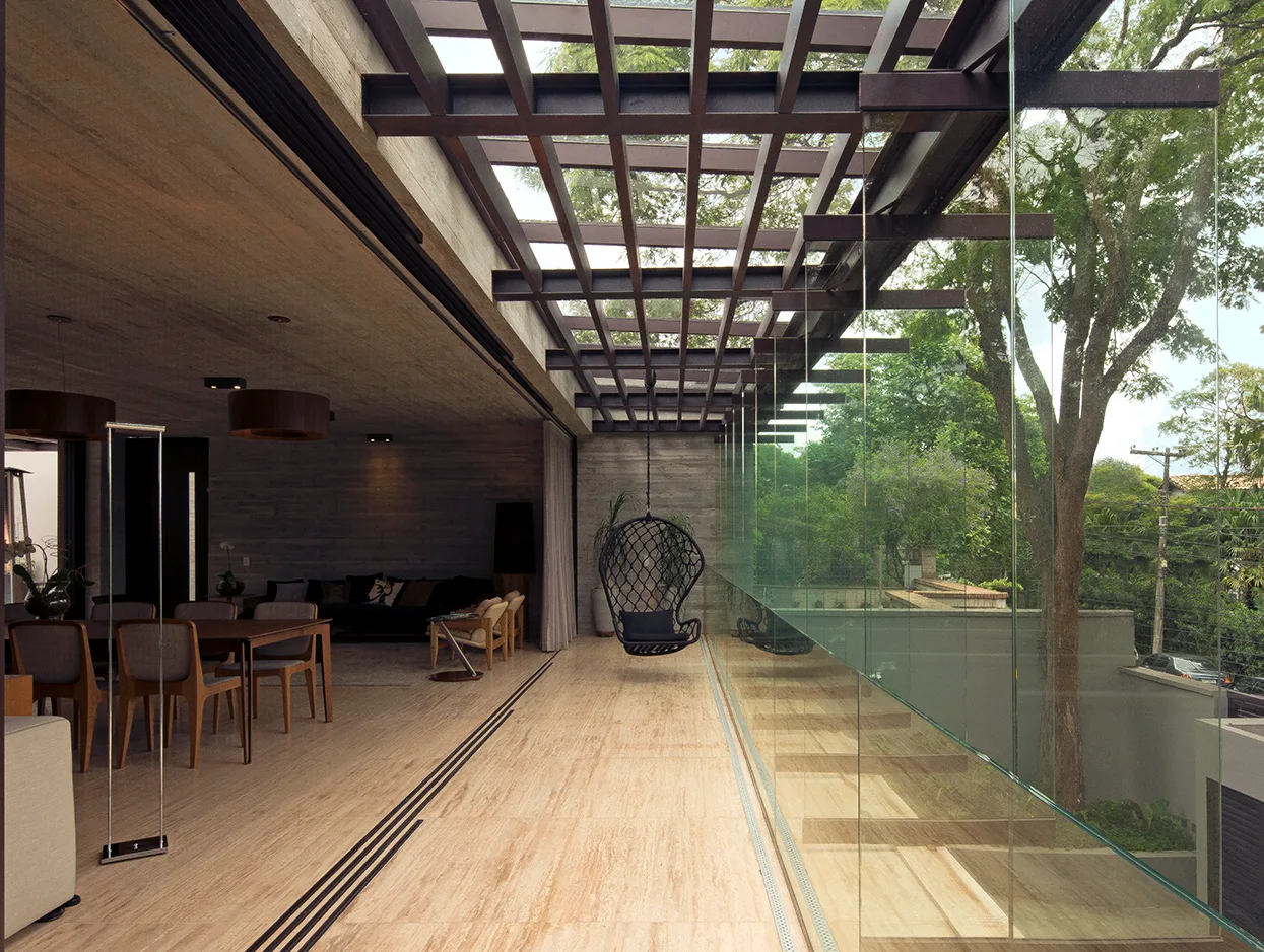 A modern indoor-outdoor living and dining area with board-formed concrete walls, beige stone floors, and a black metal pergola canopy.