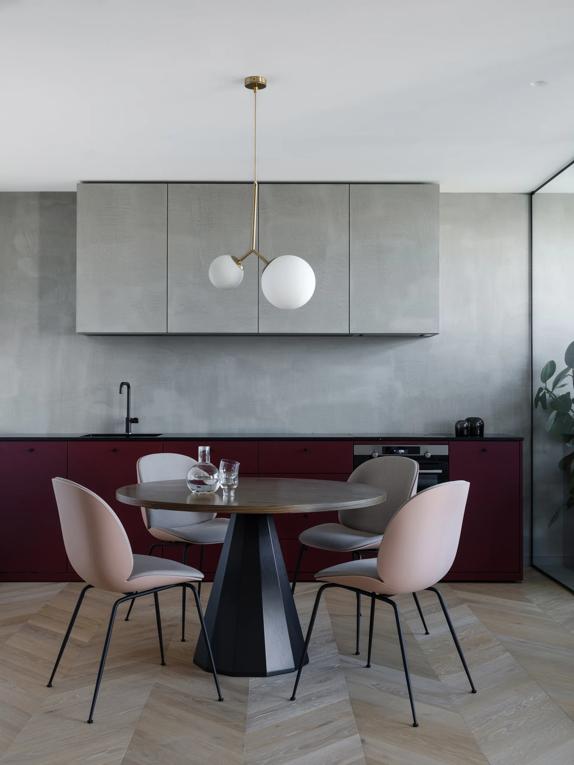 A contemporary kitchen dining area features a round wooden table, dusty rose chairs, burgundy lower cabinets, and gray concrete-style upper cabinets.