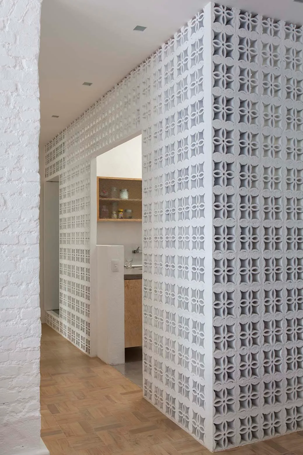 A white patterned breeze block partition wall separates rooms in a house with parquet wood floors and a painted brick wall.