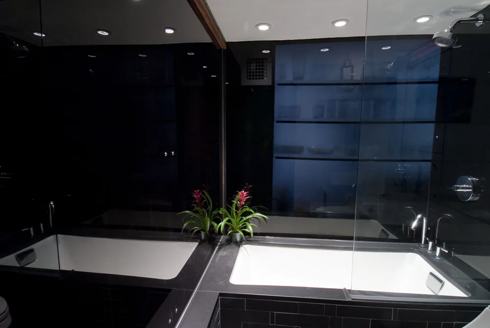 A modern bathroom featuring a white rectangular inset bathtub surrounded by dark tiles, chrome fixtures, and a large black mirrored wall.