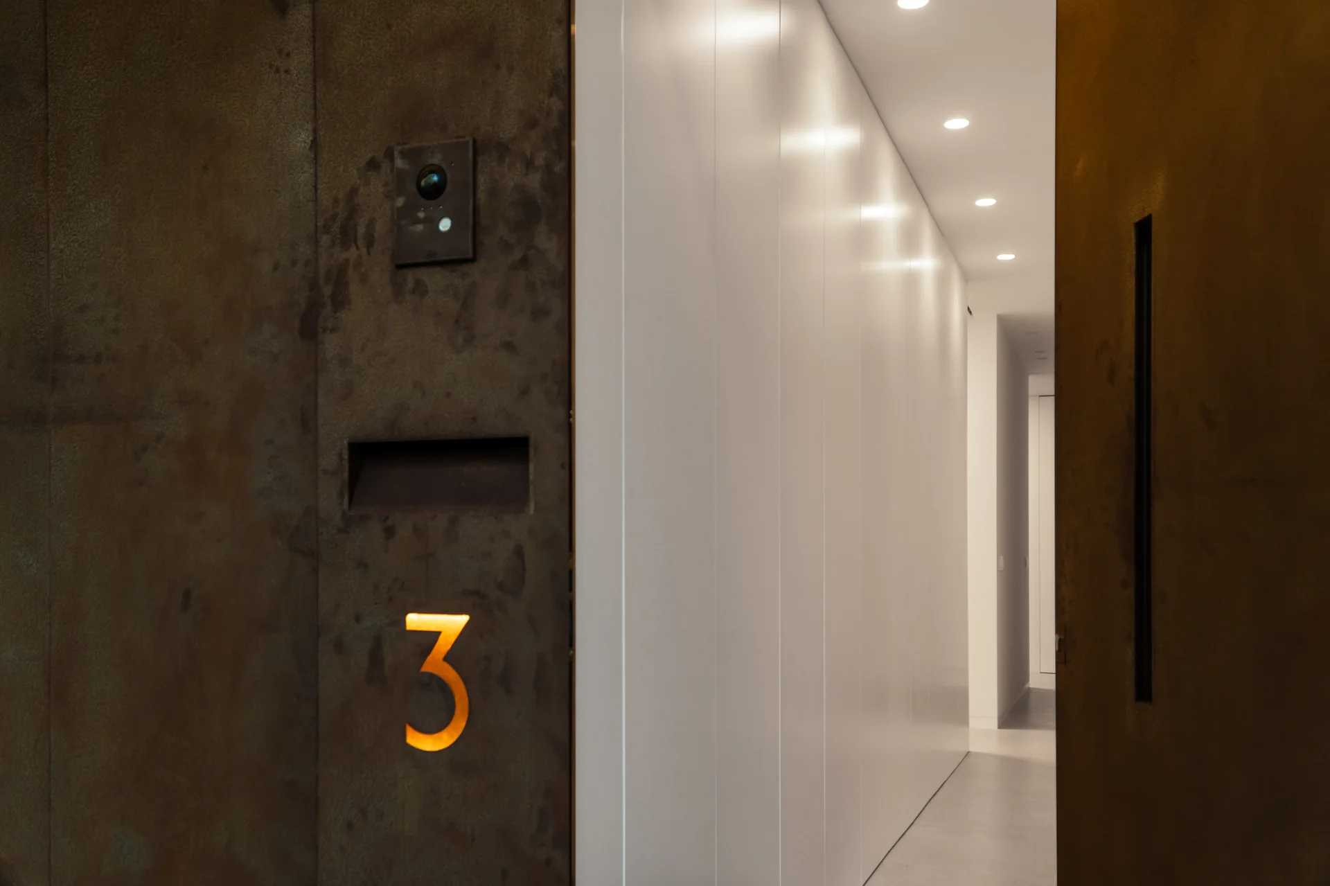 A minimalist entryway featuring a textured rust-colored metal door with an illuminated amber number three next to a bright white paneled hallway.