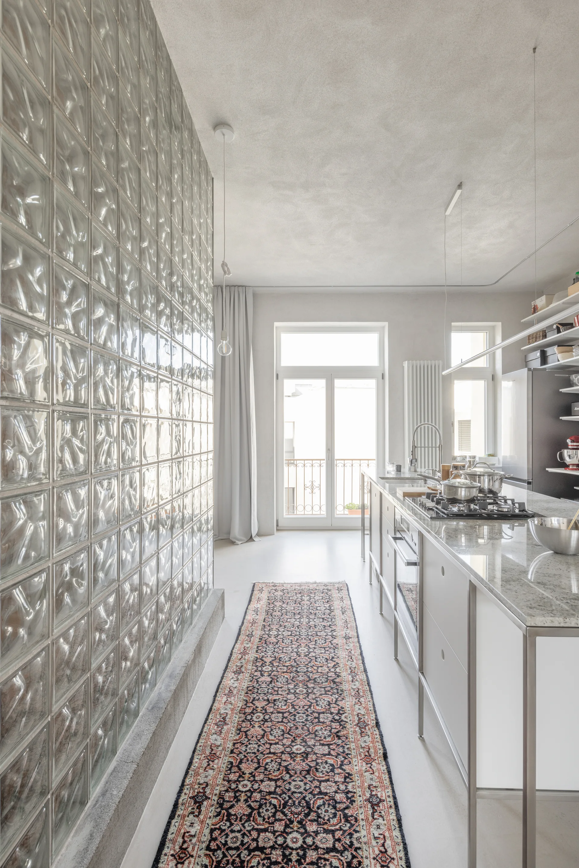 A modern kitchen featuring a floor-to-ceiling glass block wall, a stone-topped island with a cooktop, and a patterned runner rug.