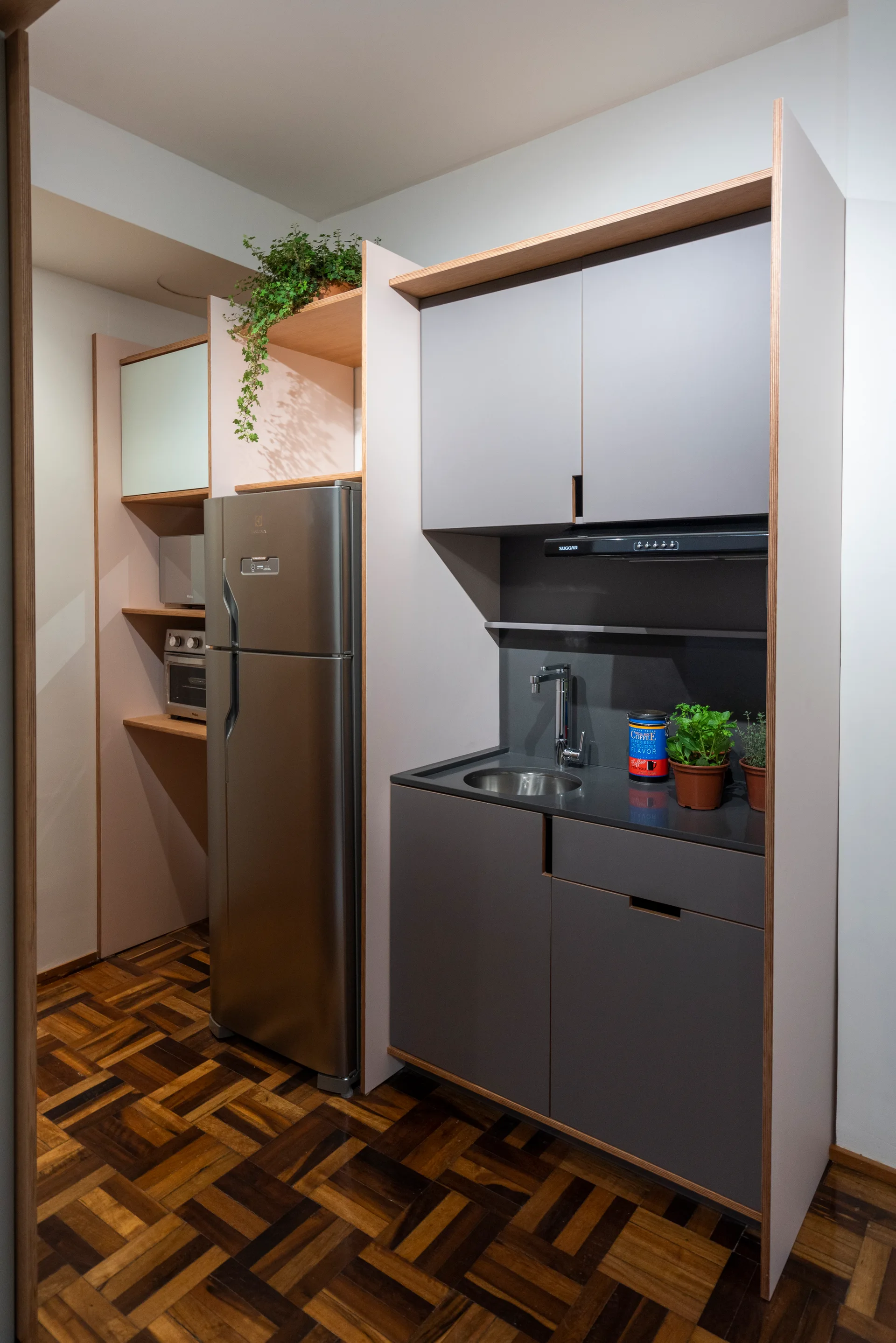 A small kitchenette features matte grey cabinets with exposed plywood trim, a stainless steel refrigerator, and dark wood parquet flooring.