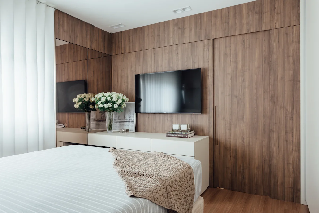 A modern bedroom with a large wood paneled feature wall, a wall-mounted flat-screen television, a low white console, and a bed in the foreground.