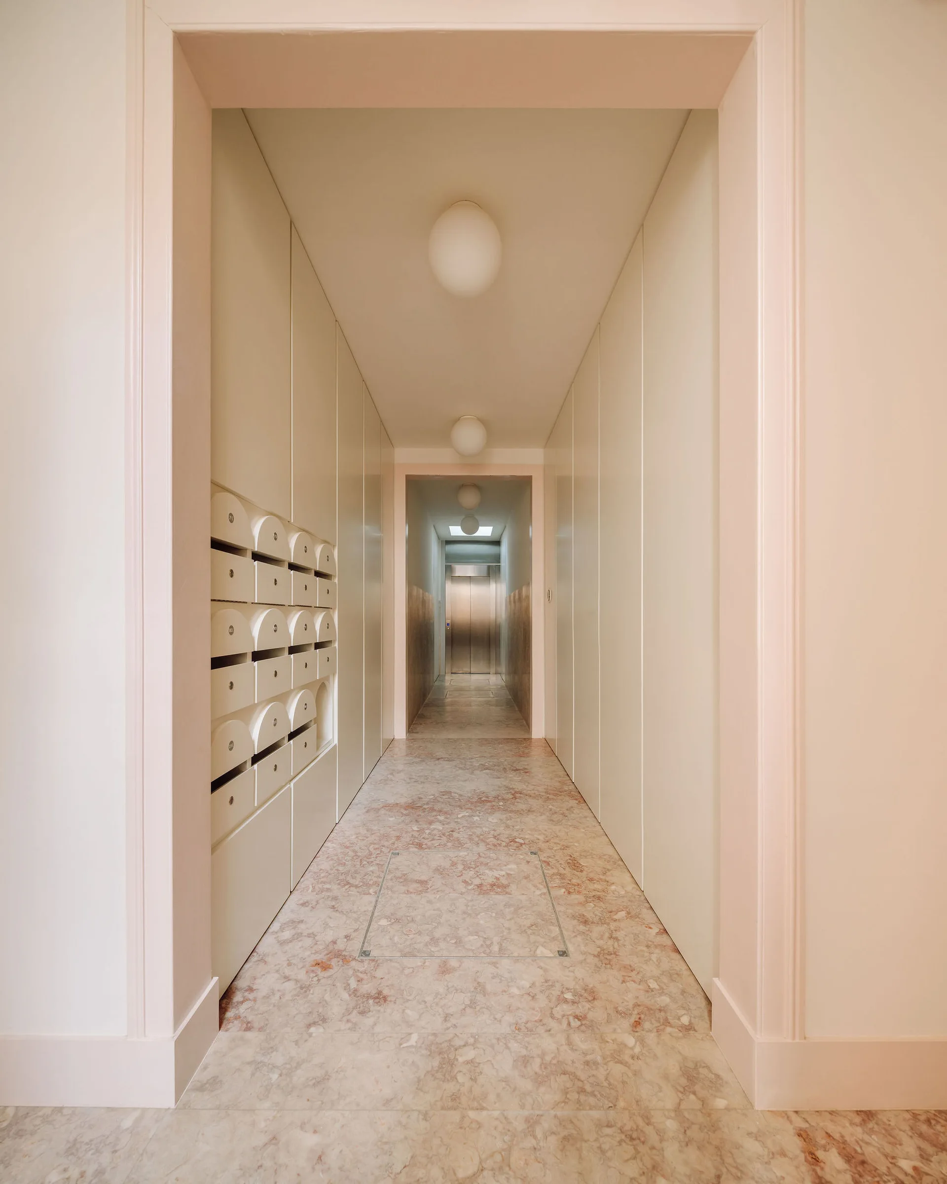A minimalist hallway features white paneled walls with integrated mailboxes, pink veined marble flooring, and globe ceiling lights leading to an elevator.