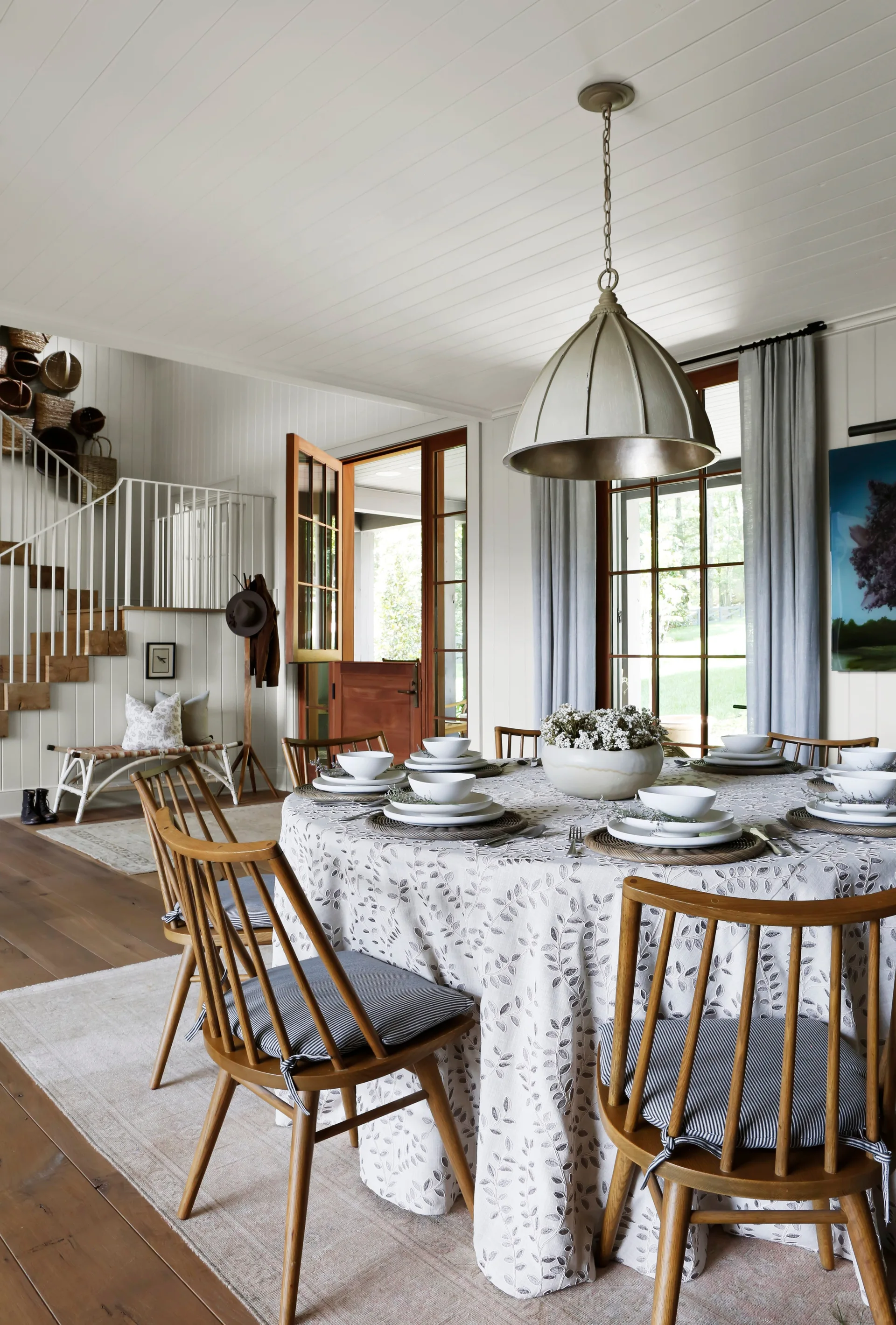A cottage-style dining room features a round table set with white dishes, wooden spindle chairs with striped cushions, and a large metal dome pendant.