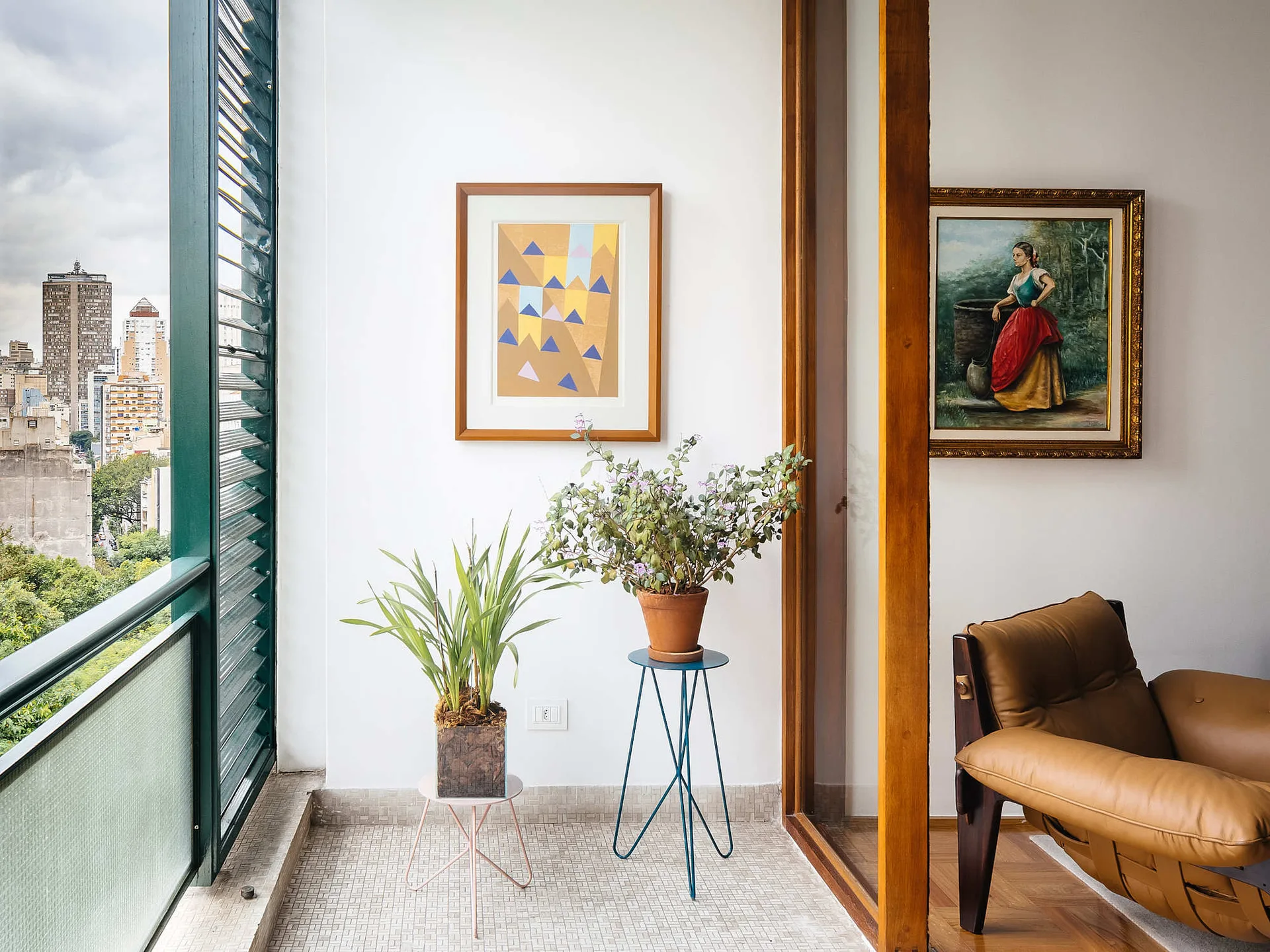 A small apartment balcony features mosaic tile flooring, potted plants on slender metal stands, framed artwork on a white wall, and a view of a city.