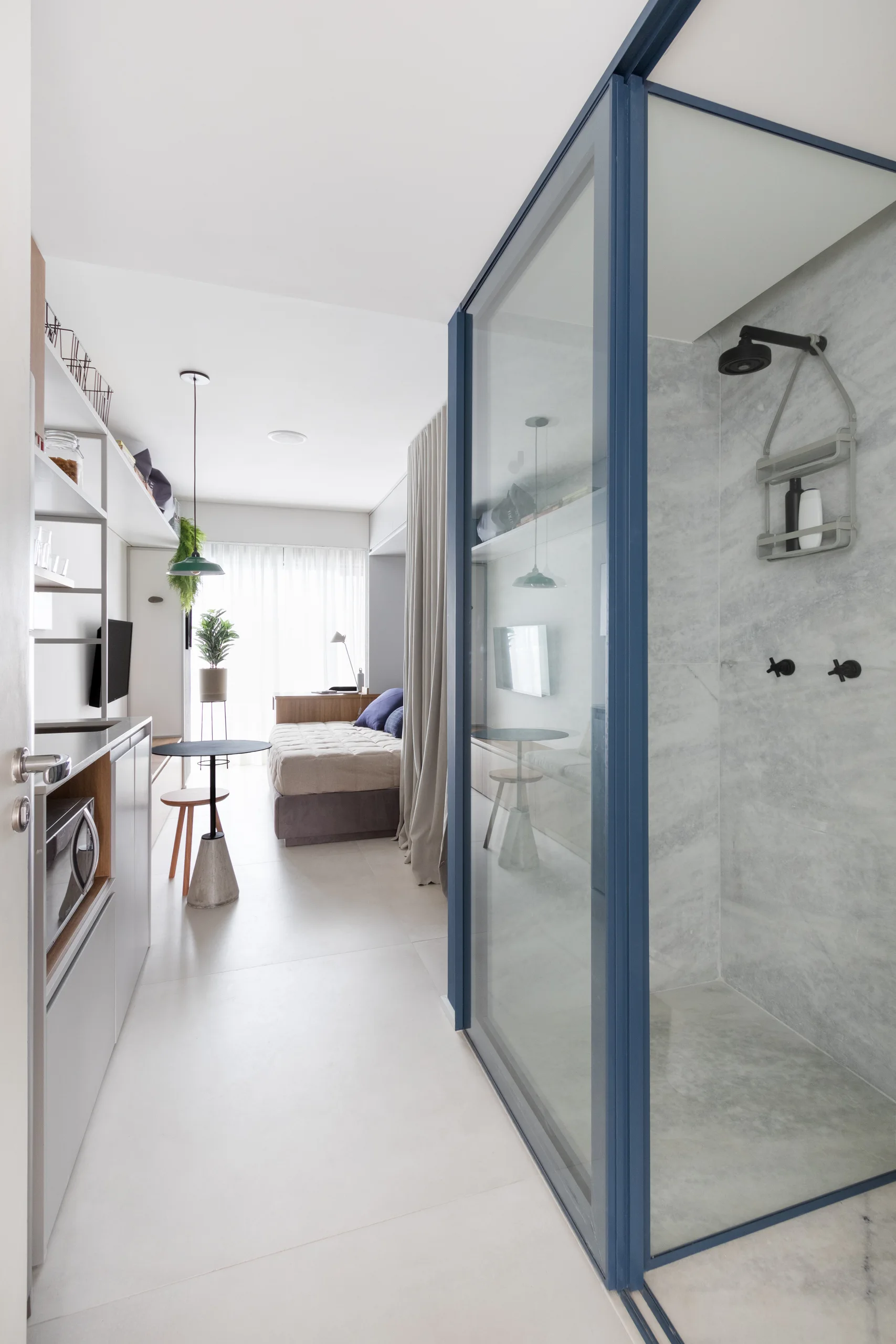A studio apartment corridor features a blue-framed glass shower enclosure next to a small dining area and a daybed in the background.