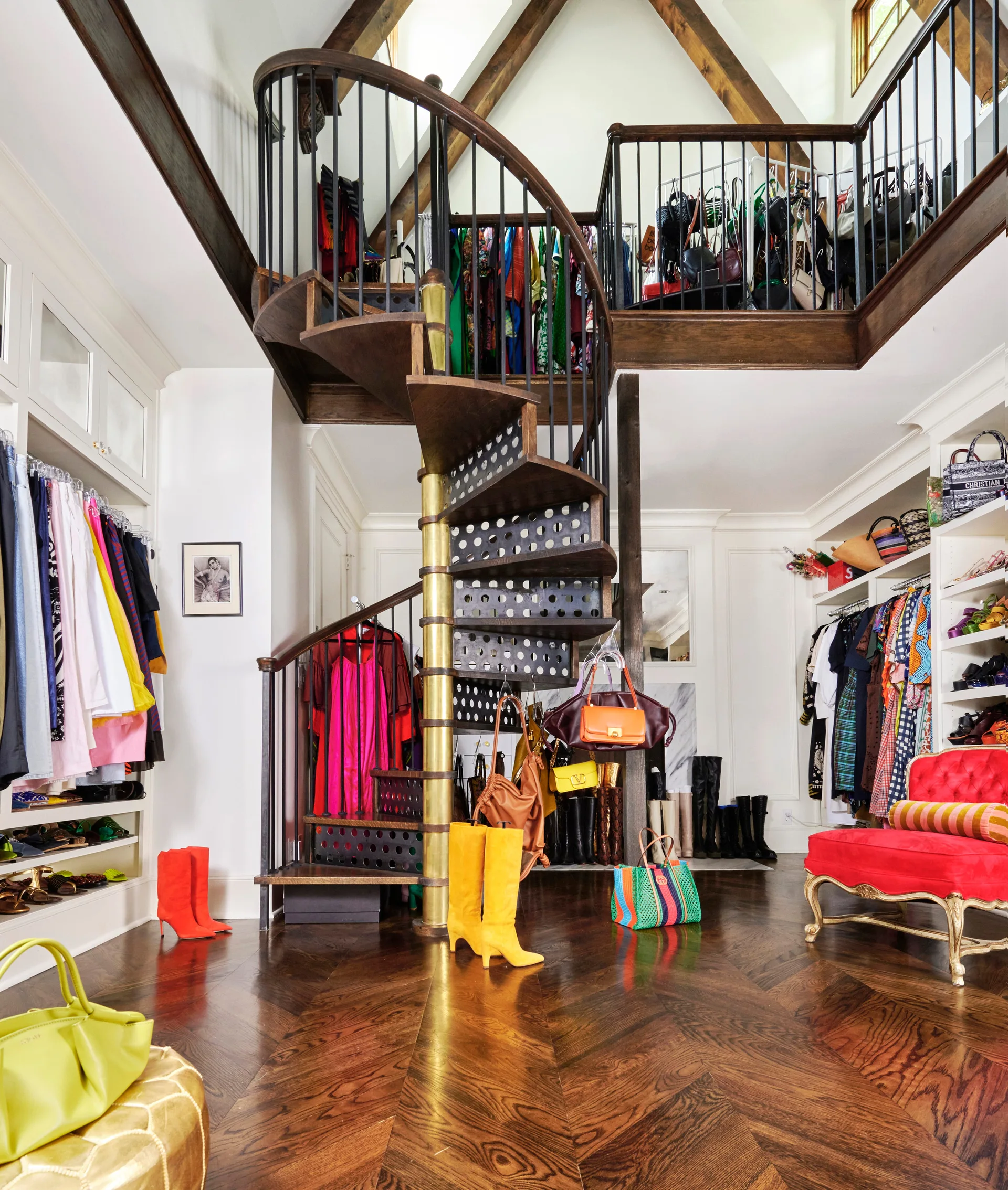A multi-level walk-in closet featuring a dark wood spiral staircase, herringbone flooring, white built-in shelving, and a red armchair.
