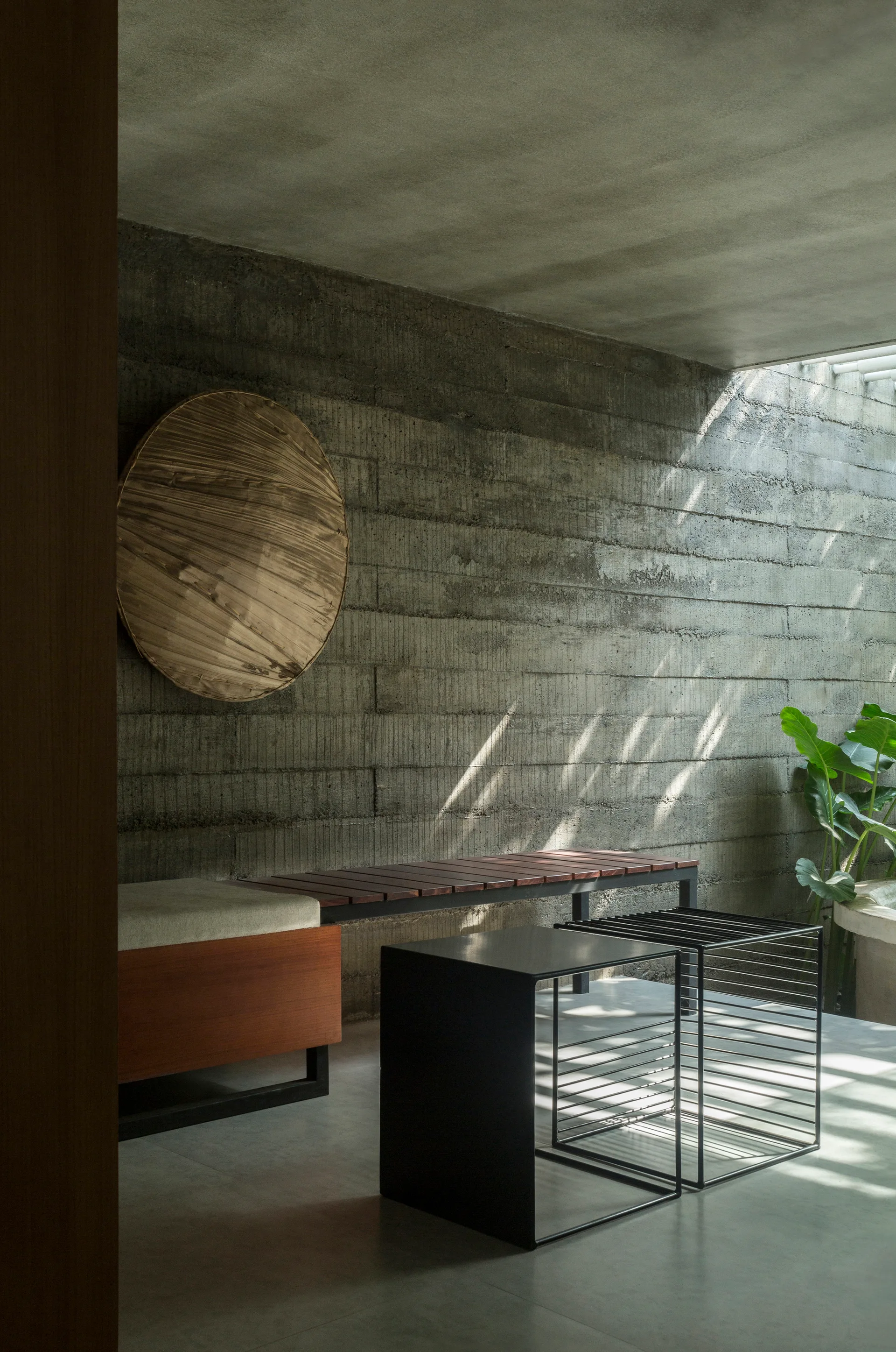 A minimalist entryway features a board-formed concrete wall, a wood slatted bench with a cushion, black metal tables, and sunbeam shadows.